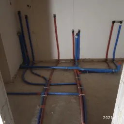 Surowy stan: Widok na podłogę z częściowo ułożonymi, niebieskimi i czerwonymi rurami instalacji wodnej i grzewczej w pomieszczeniu w trakcie remontu, z widocznymi fragmentami ścian w stanie...