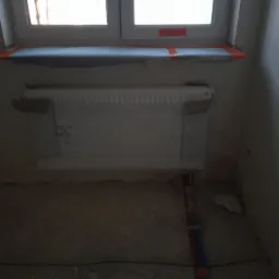 Grzejnik żeberkowy zamontowany pod oknem, zabezpieczony taśmą malarską podczas prac wykończeniowych, z widocznymi rurami instalacji grzewczej w podłodze.