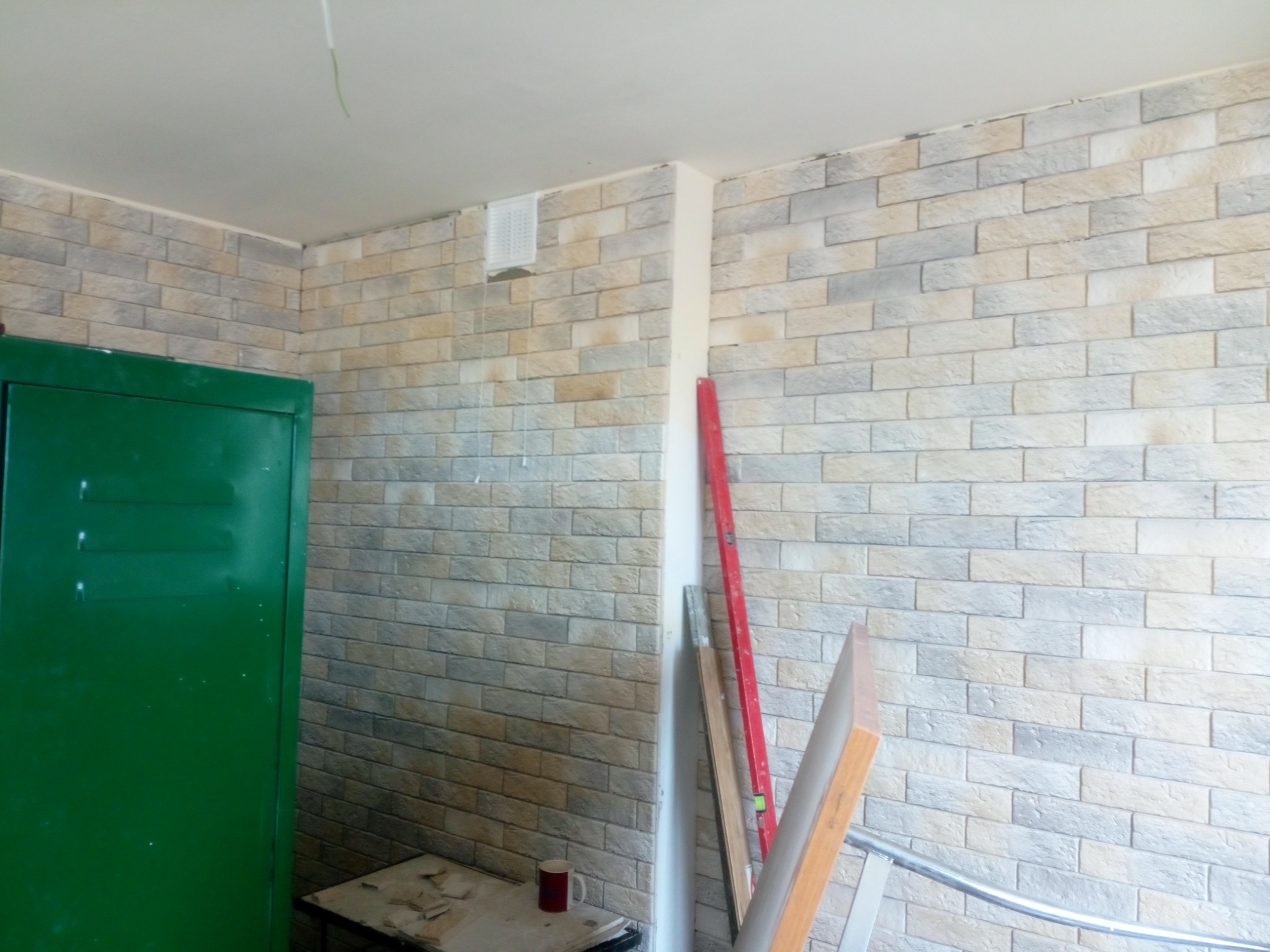 Ściana wyłożona dekoracyjną cegłą w odcieniach szarości i beżu, z widoczną kratką wentylacyjną i pionową szczeliną, obok stoi zielona szafa i leżą narzędzia.