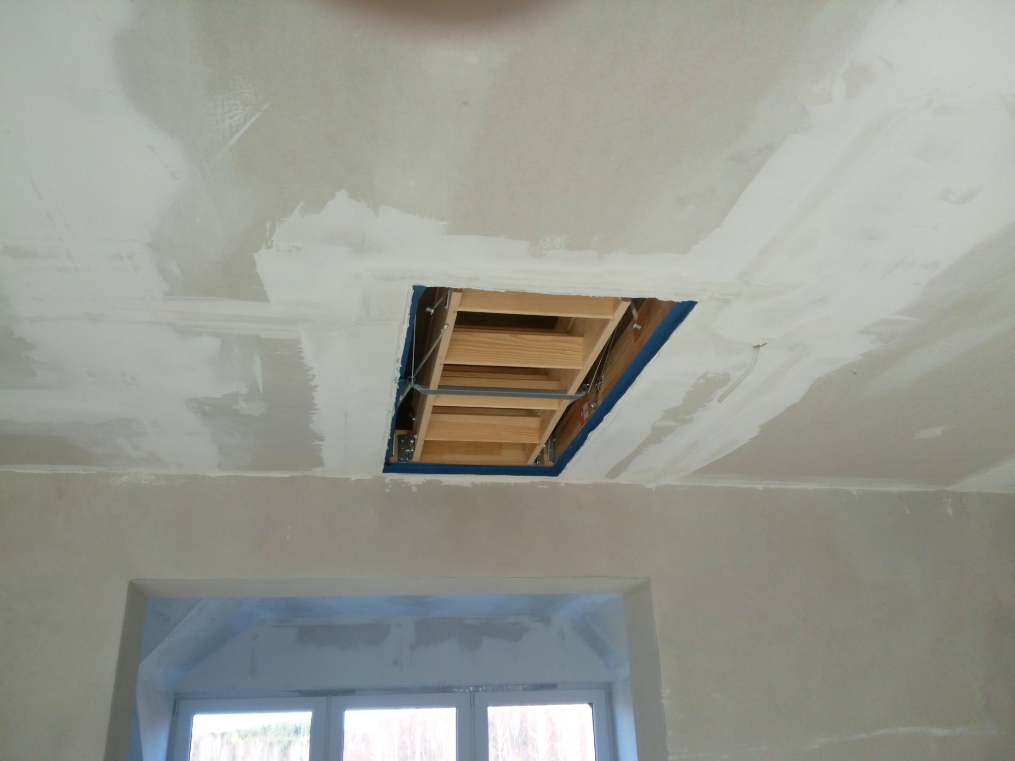 Sufit z otworem na schody strychowe w trakcie montażu, widoczne łatki gipsowe i drewniana konstrukcja schodów, w tle okno.