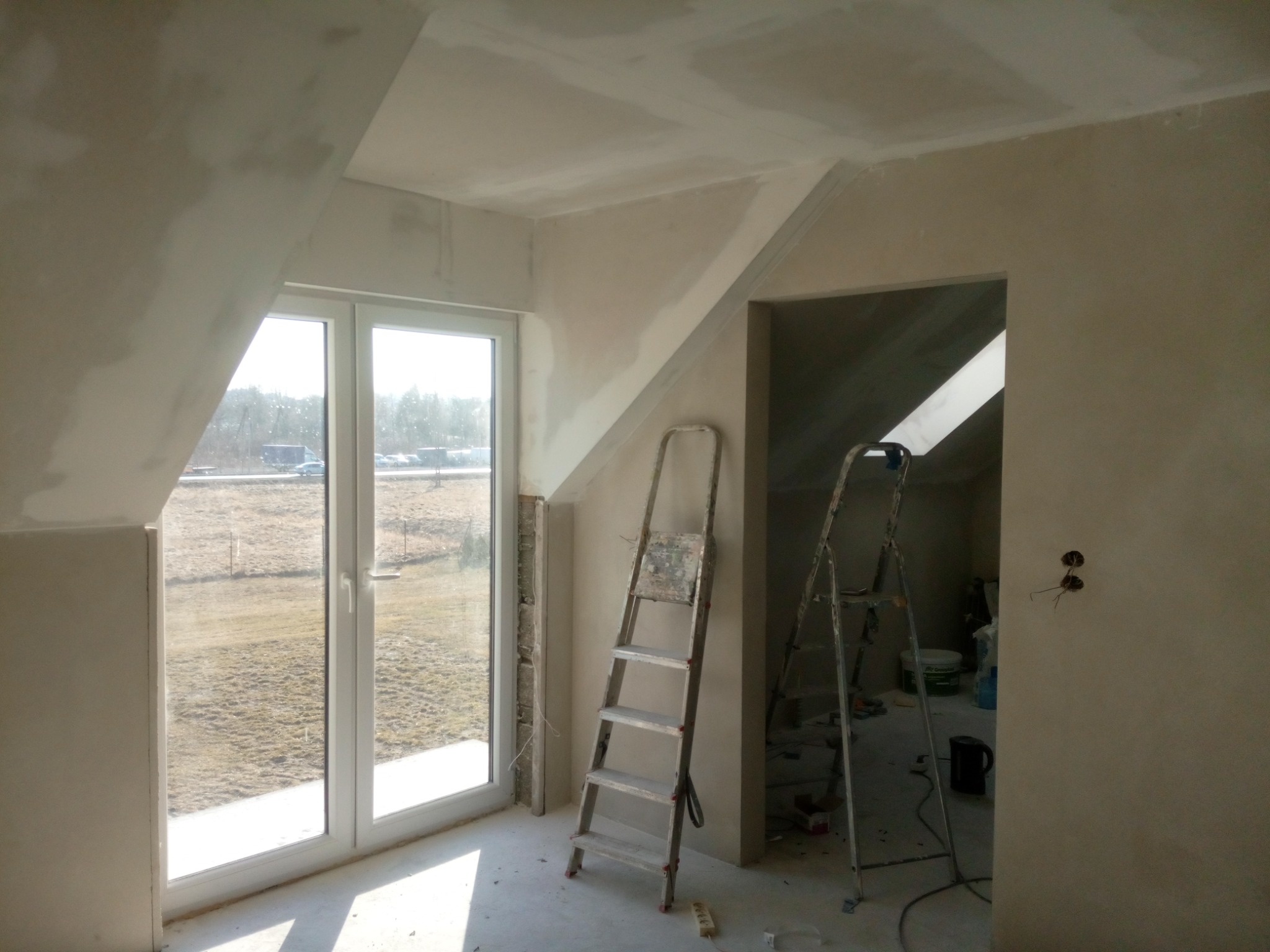 Pokój na poddaszu w trakcie prac wykończeniowych, widoczne dwie drabiny malarskie, okno dachowe i ściany po wstępnym nałożeniu gładzi.