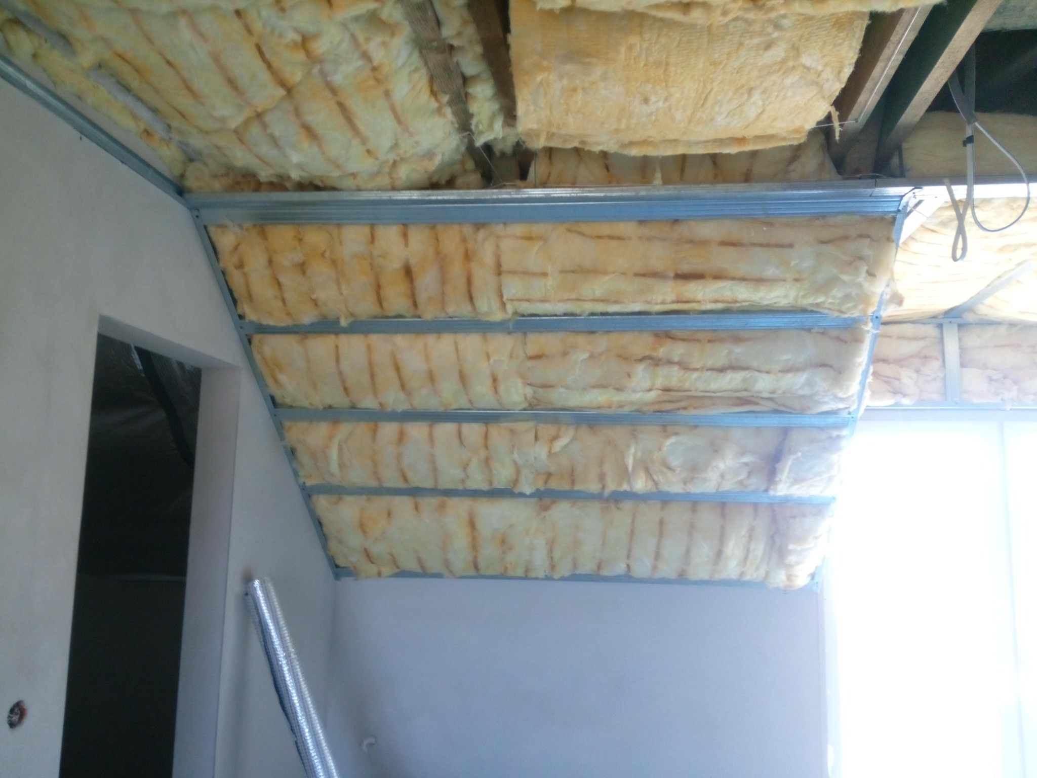 Izolacja z wełny mineralnej zamontowana w metalowym stelażu poddasza, widoczne elementy konstrukcyjne i fragment ściany z otworem drzwiowym.