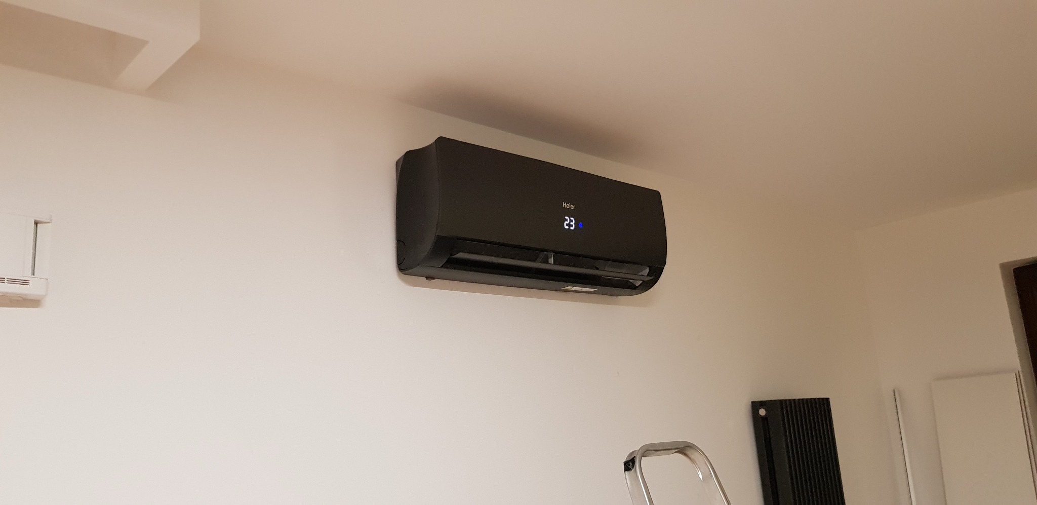 Czarny klimatyzator Haier zamontowany na białej ścianie w domu, widoczny fragment drabiny i grzejnika.