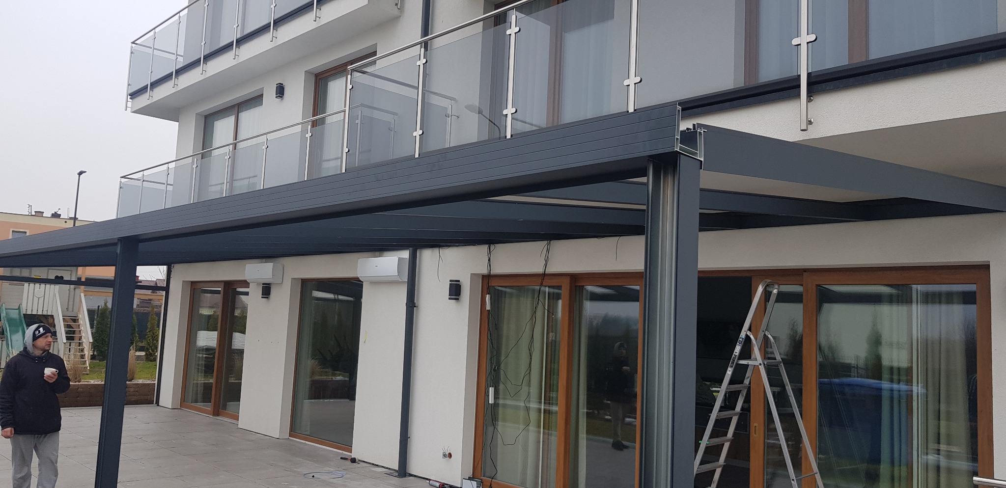 Nowoczesna pergola z ciemnoszarej konstrukcji metalowej przy białym domu w Wyszkowie. Widoczne klimatyzatory na ścianie, drabina oparta o okno i mężczyzna z kubkiem w tle.