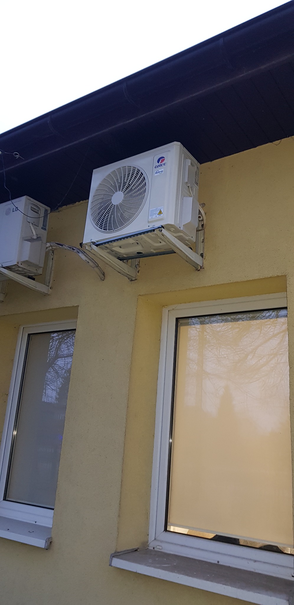 Dwa klimatyzatory zewnętrzne, marki Gree i LG, zamontowane na elewacji budynku z żółtym tynkiem, widoczne okna z białymi ramami.