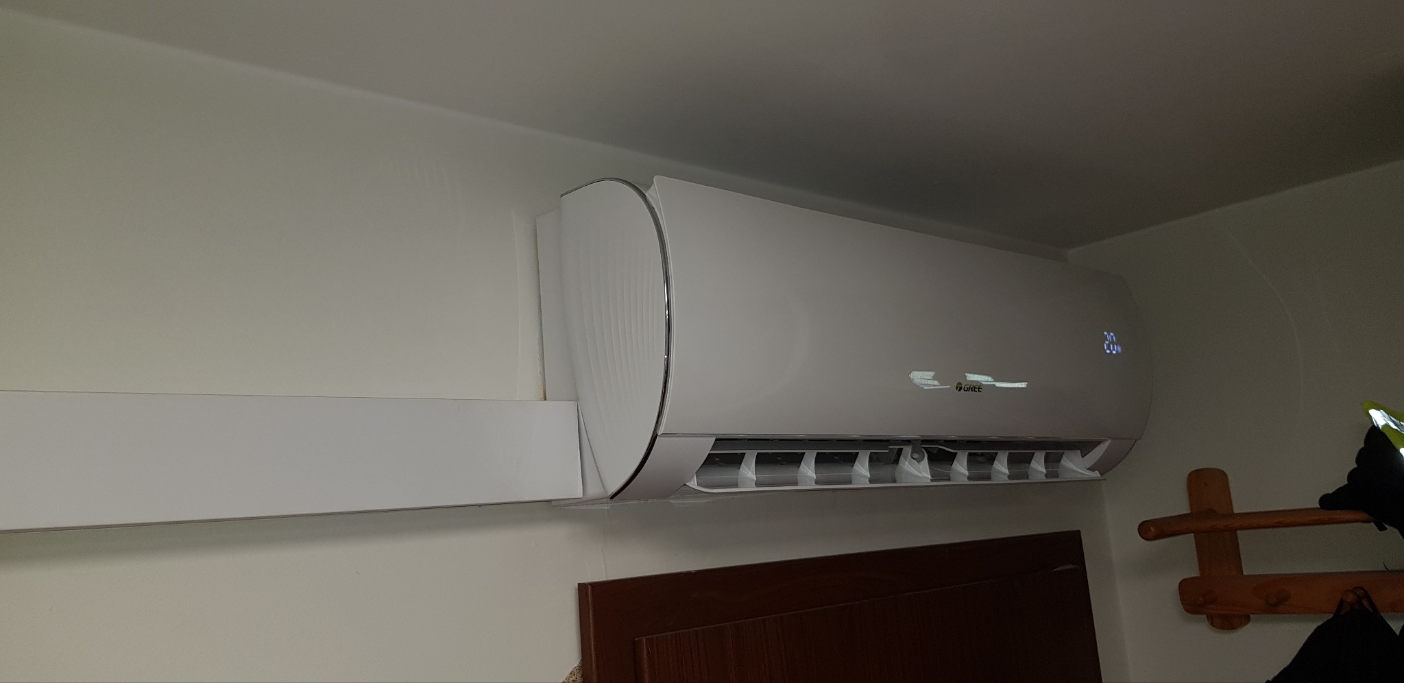 Biały klimatyzator marki Gree zamontowany na ścianie w pomieszczeniu, widoczny wyświetlacz temperatury 20 stopni oraz wieszak na ubrania w tle.