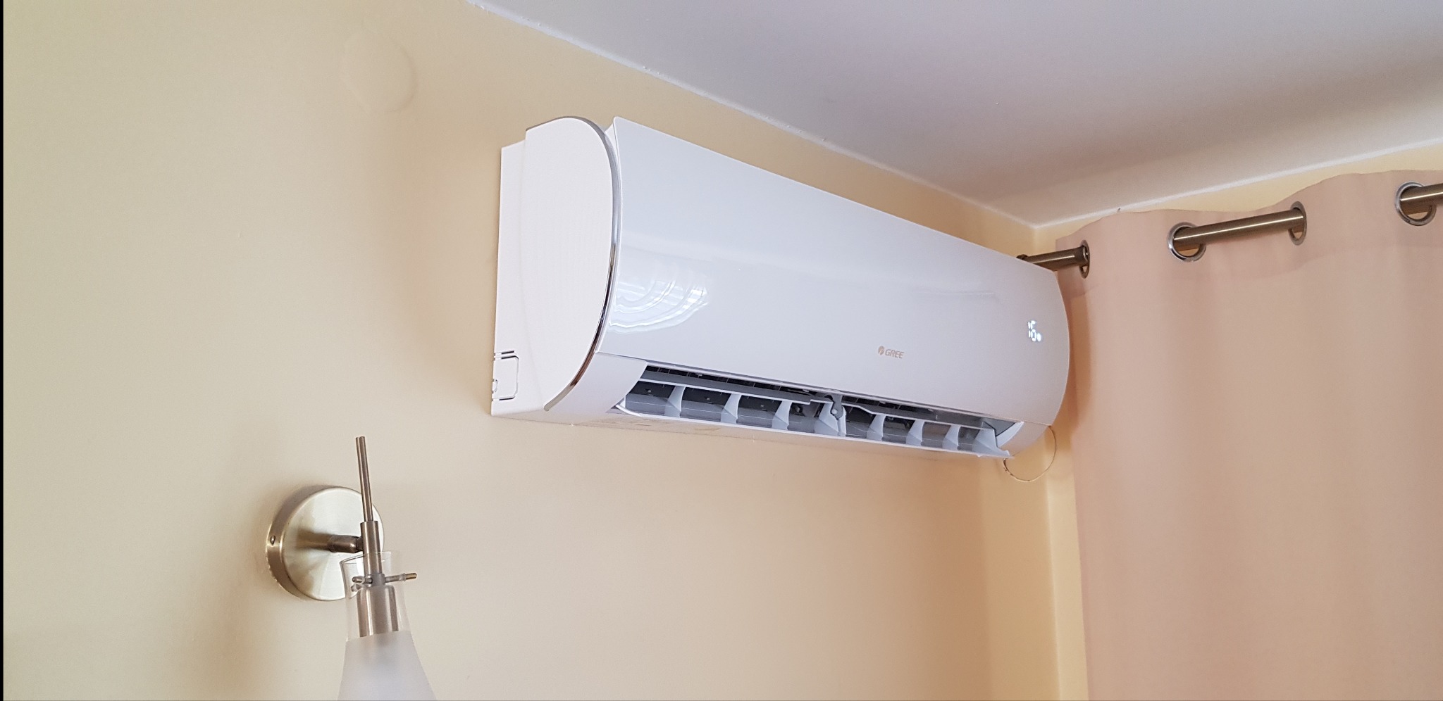Biała klimatyzacja marki Gree zamontowana na jasnej ścianie w pokoju, widoczna lampka nocna i zasłona.
