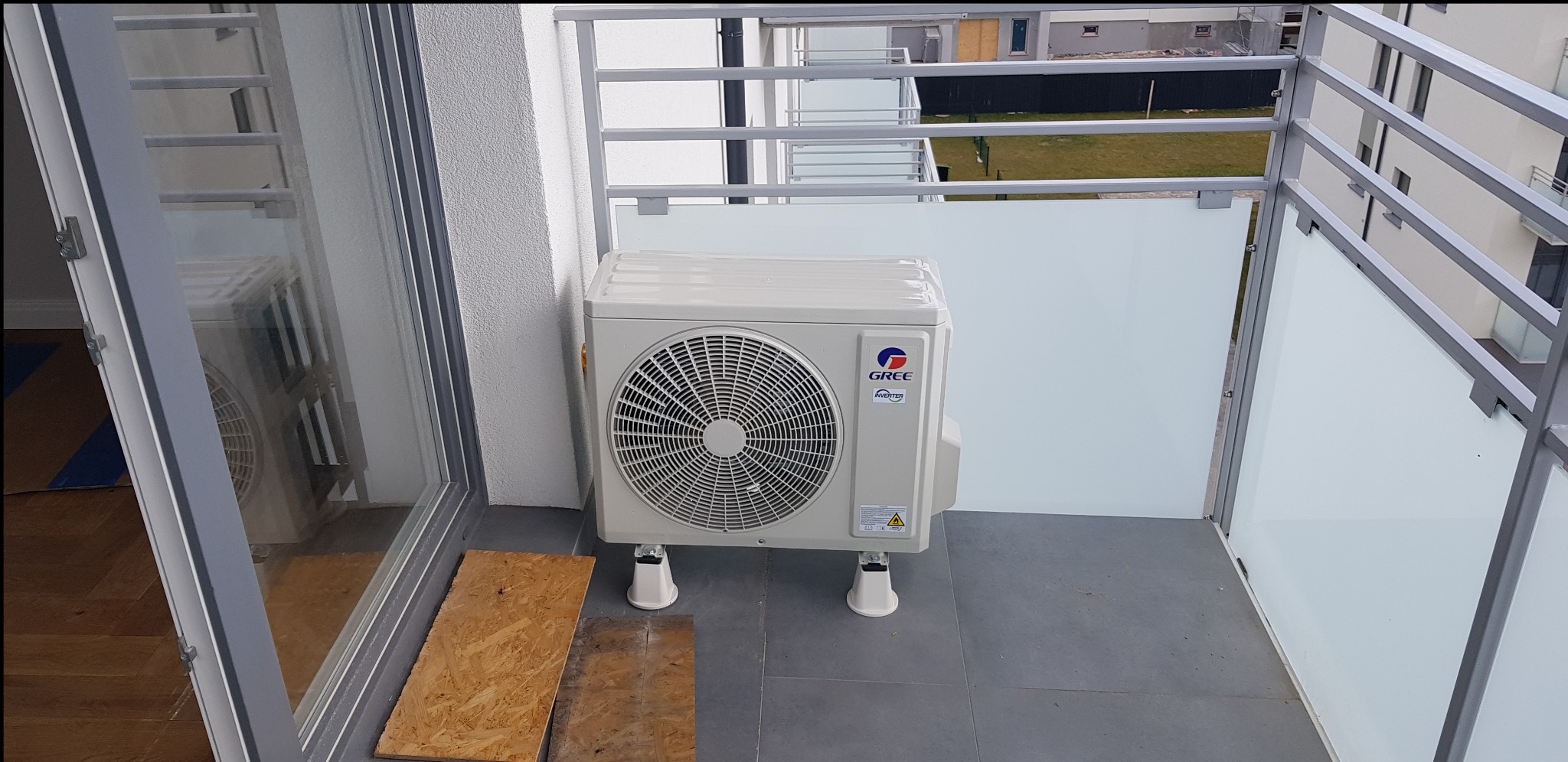 Jednostka zewnętrzna klimatyzatora marki Gree umieszczona na balkonie z szarymi płytkami, zabezpieczonym metalową balustradą z mlecznym szkłem.