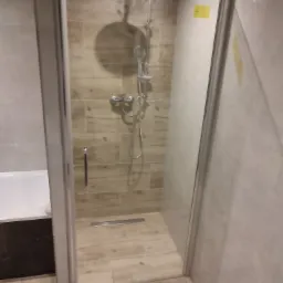 Szklana kabina prysznicowa z odpływem liniowym, wykończona płytkami imitującymi drewno, z widocznym fragmentem wanny obok.