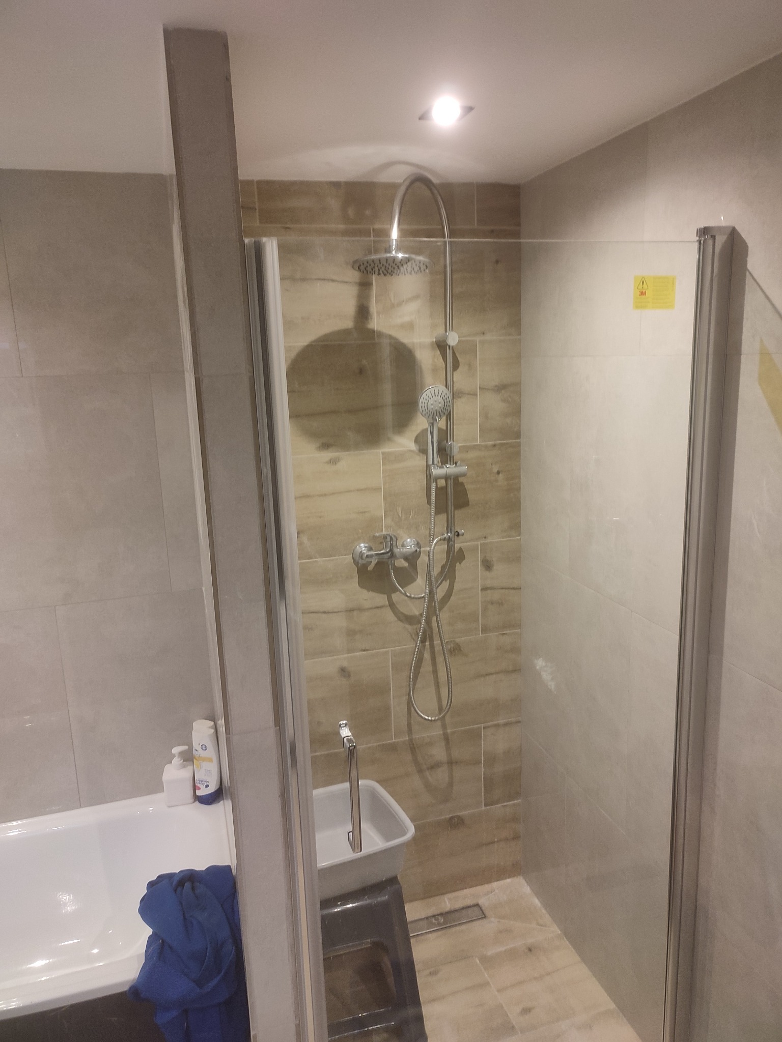 Nowoczesna kabina prysznicowa z deszczownicą i panelami ściennymi imitującymi drewno, widoczna umywalka na podeście i fragment wanny z niebieskim ręcznikiem, oświetlenie punktowe sufitowe.