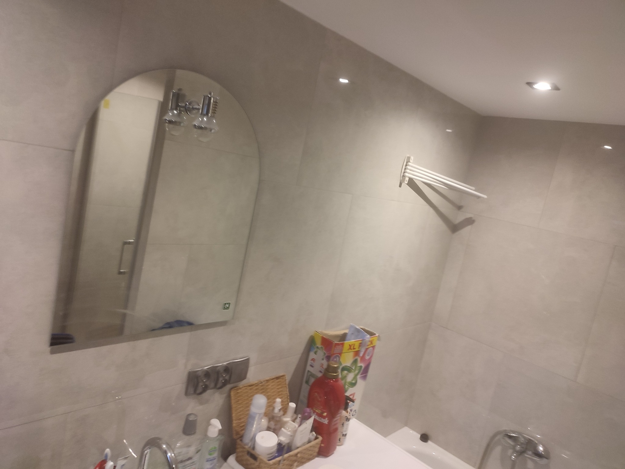 Jasna łazienka z beżowymi płytkami, owalne lustro z oświetleniem, biały grzejnik drabinkowy na ścianie, koszyk z kosmetykami na blacie.