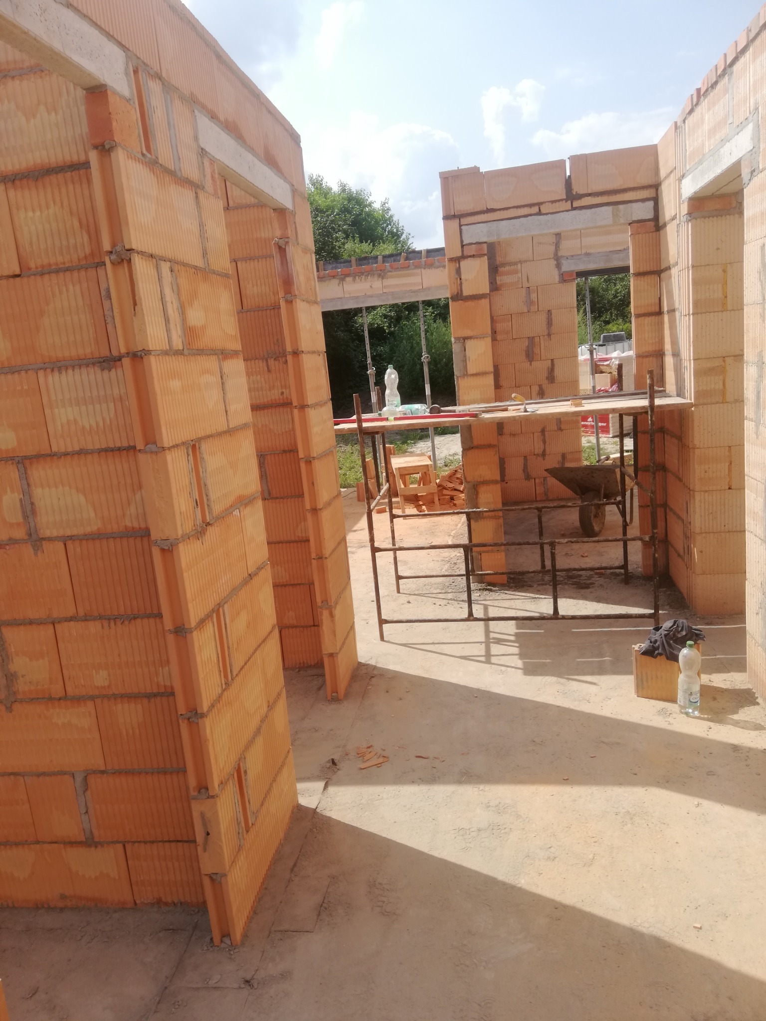 Wnętrze budynku w trakcie budowy, widoczne ściany z czerwonej cegły ceramicznej, rusztowanie robocze, taczka i butelka wody mineralnej.