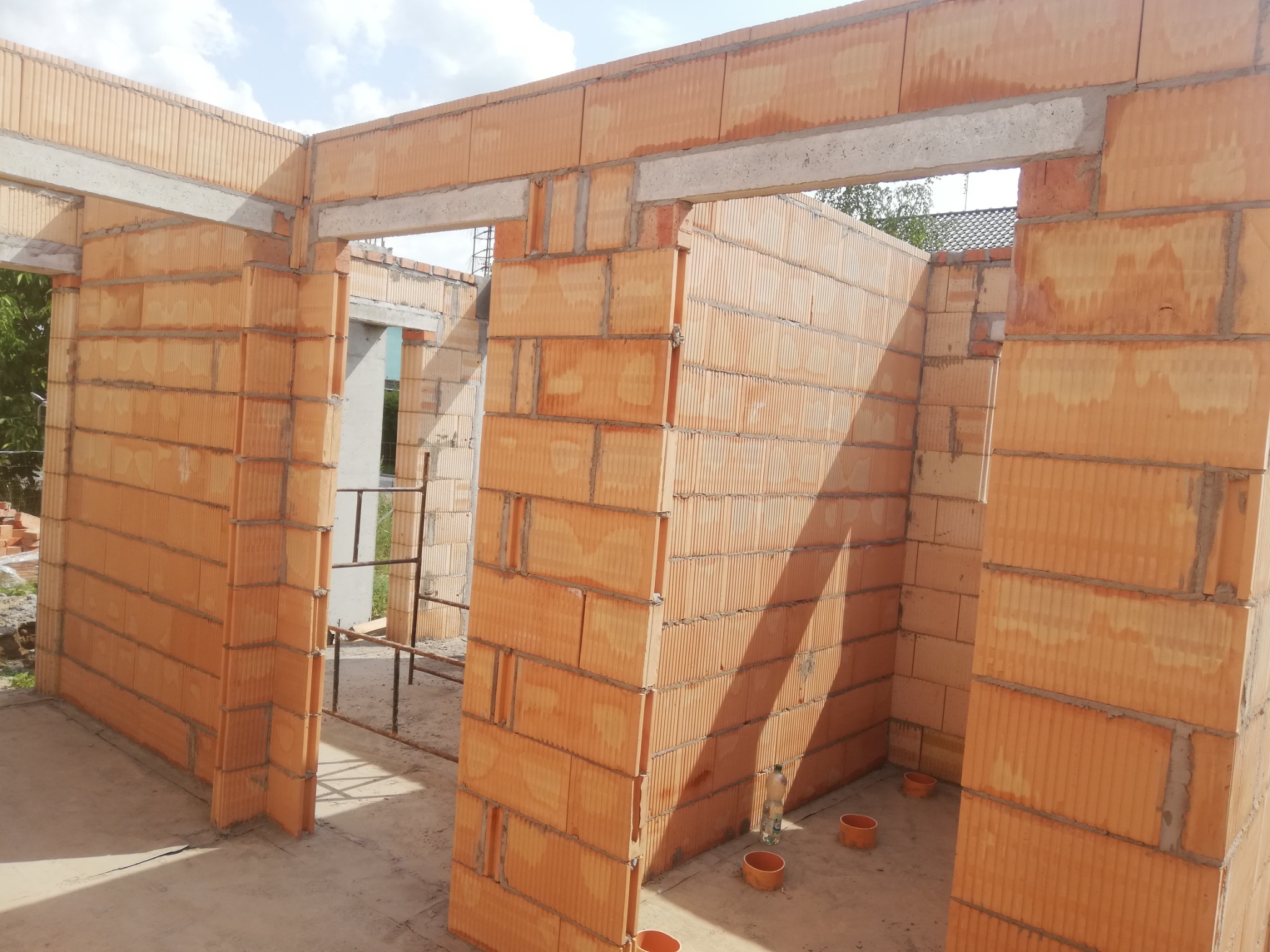 Wnętrze domu w trakcie budowy: ściany z pomarańczowych pustaków ceramicznych, betonowe nadproża, widoczne rusztowanie i butelka wody na podłodze.