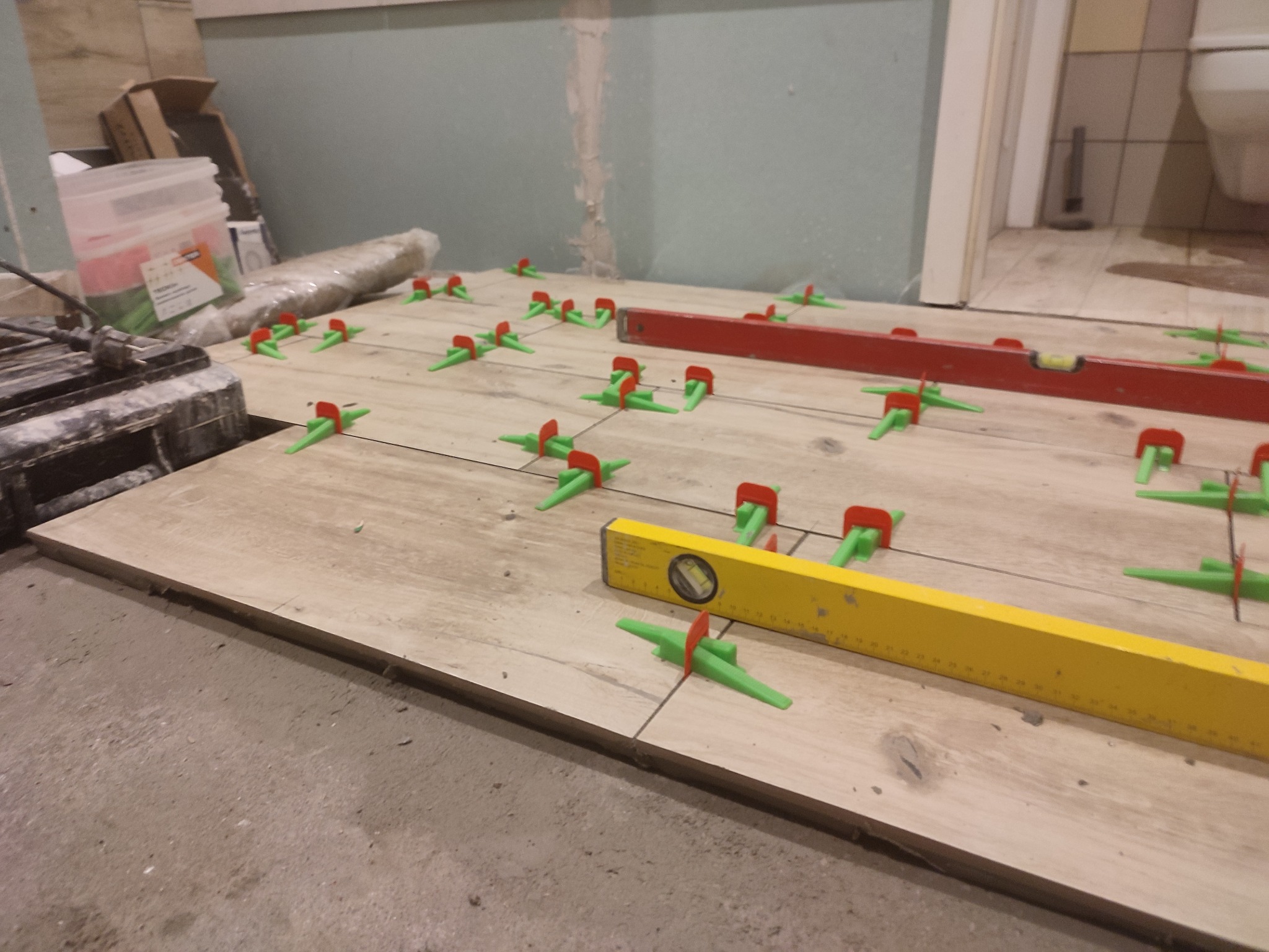 Układanie płytek podłogowych w łazience z użyciem systemu poziomowania, widoczne poziomice i paczka zaprawy klejowej w tle.