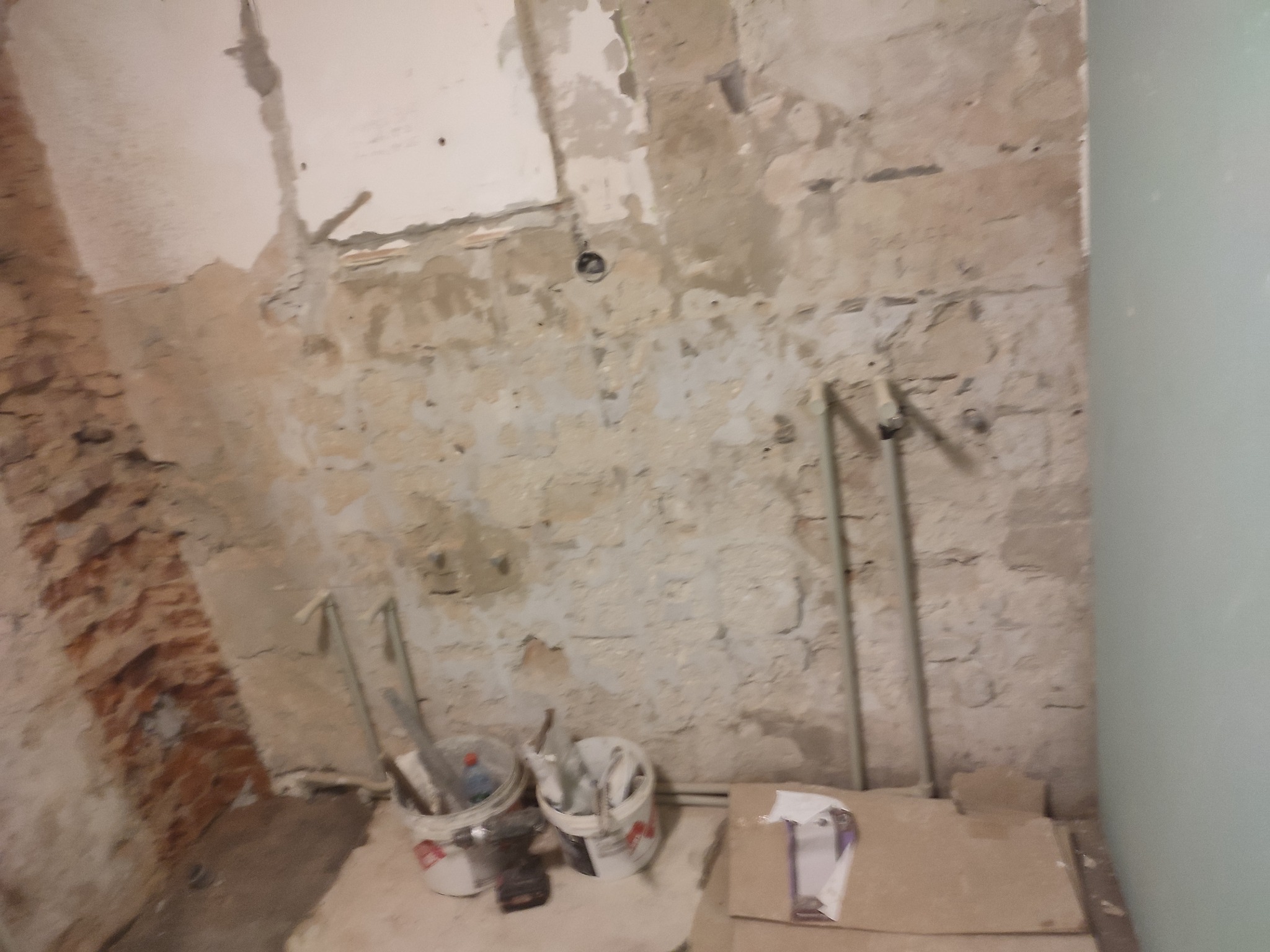 Ściana w trakcie remontu z odsłoniętą cegłą, fragmentem płyty, instalacją hydrauliczną i dwoma wiadrami z narzędziami.