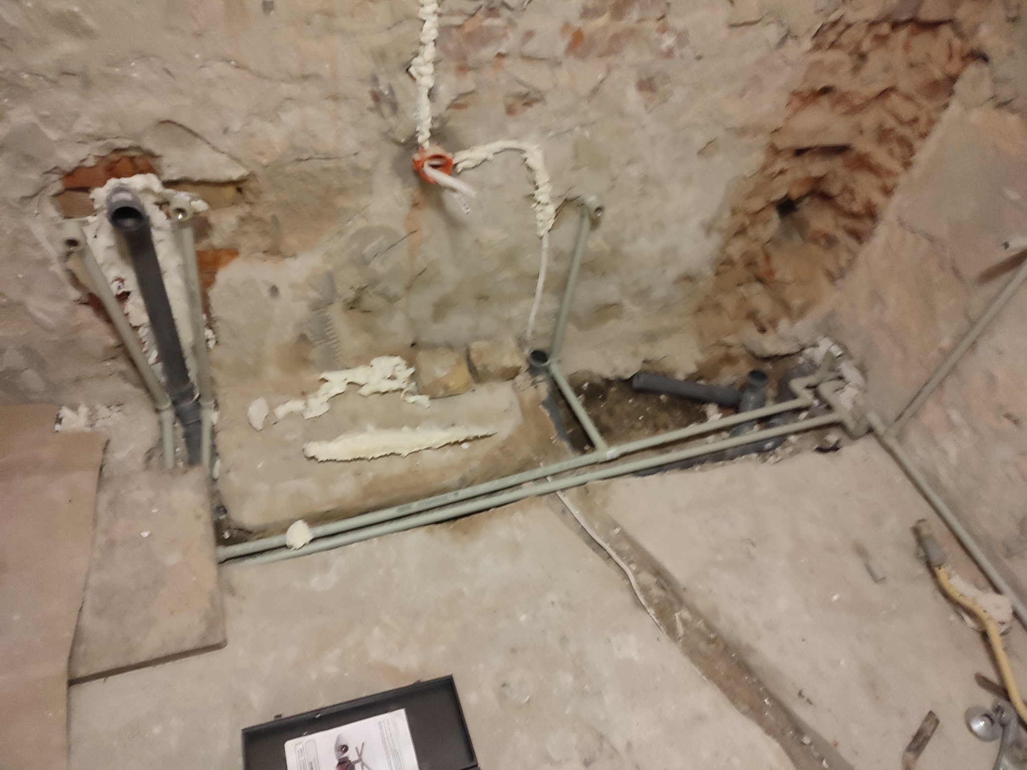 Surowy stan łazienki w trakcie prac remontowych: widoczne rury kanalizacyjne przytwierdzone do ściany pianką montażową, fragmenty odsłoniętej cegły i narzędzia na podłodze.