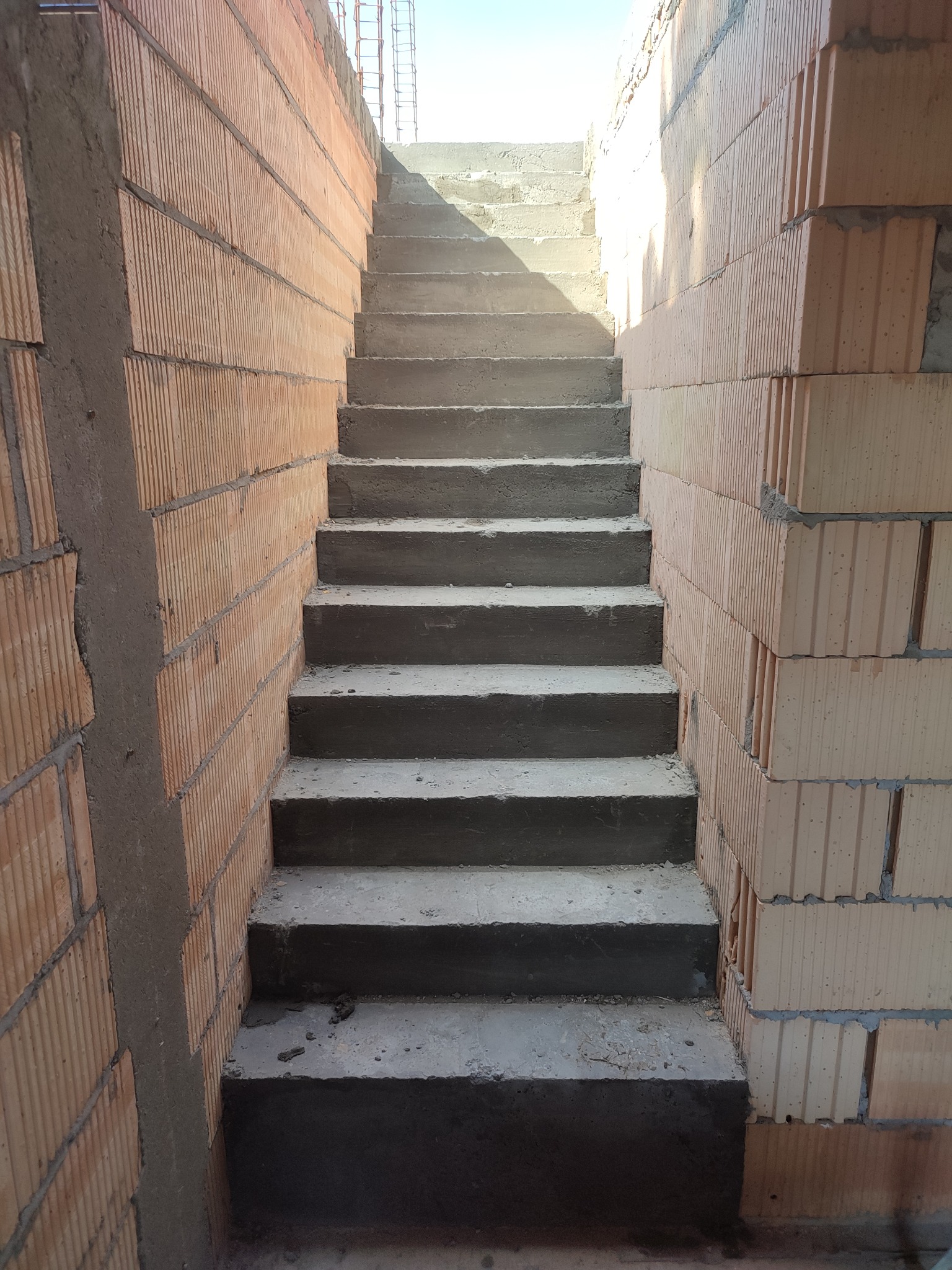 Surowe betonowe schody w trakcie budowy, otoczone ścianami z cegły ceramicznej dziurawki, widok z dołu w kierunku światła.