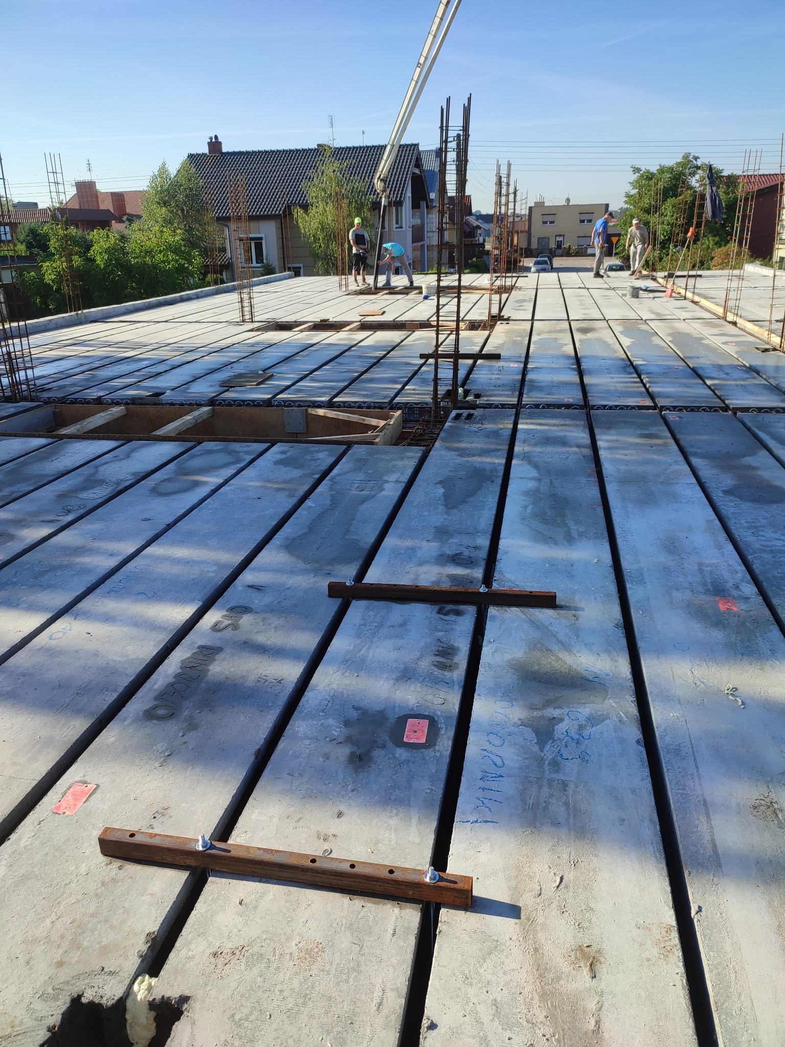 Wylewanie betonu na strop Teriva z widocznymi pustakami, zbrojeniami i konstrukcją wsporczą, w tle budynki mieszkalne i pracownicy budowlani.