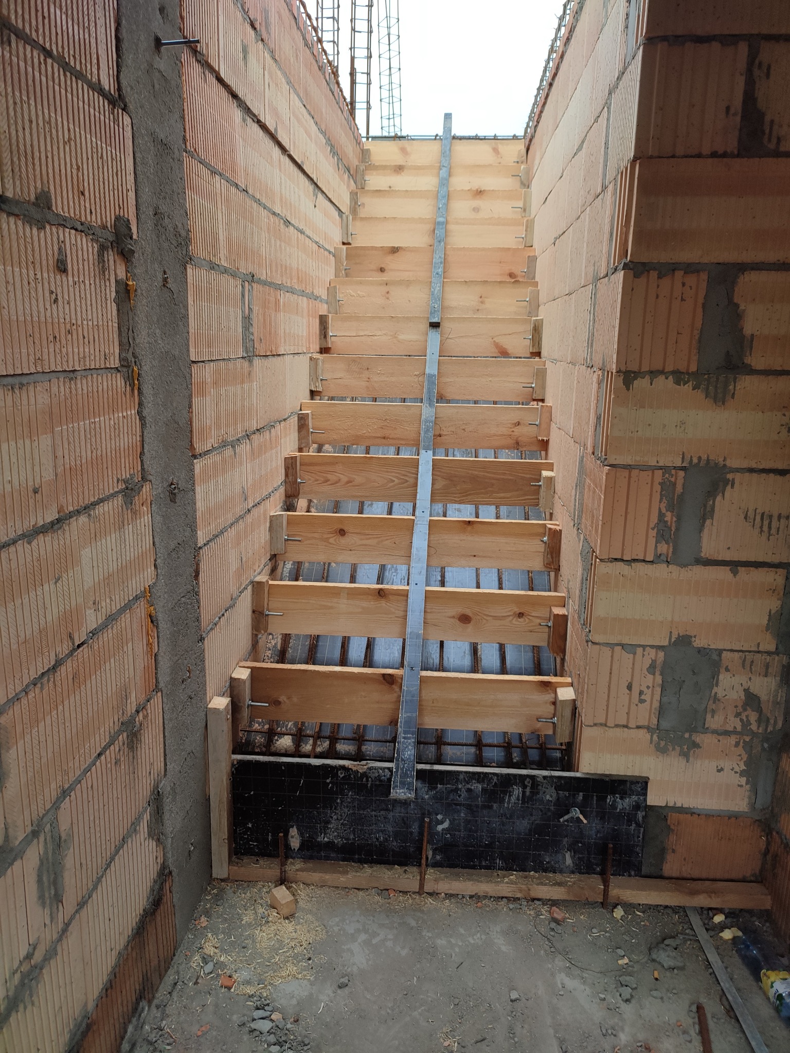 Wewnętrzne schody w trakcie budowy, szalunek z desek pomiędzy ścianami z czerwonej cegły ceramicznej, widoczne zbrojenie.