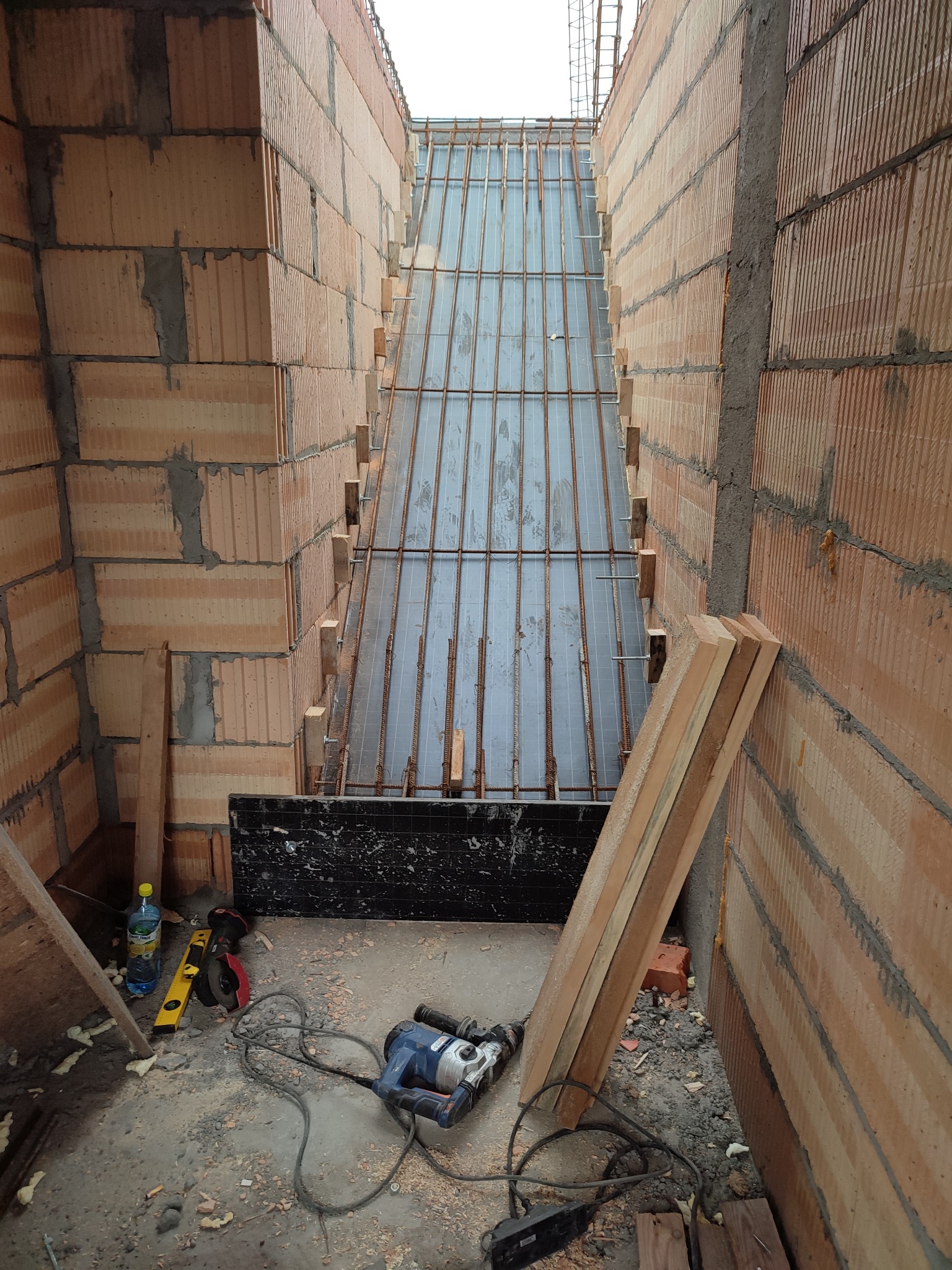 Wnętrze budowy z cegieł, widoczne zbrojenie i deskowanie pod wylewkę. Na podłodze narzędzia budowlane, poziomica, butelka z wodą i resztki materiałów budowlanych.