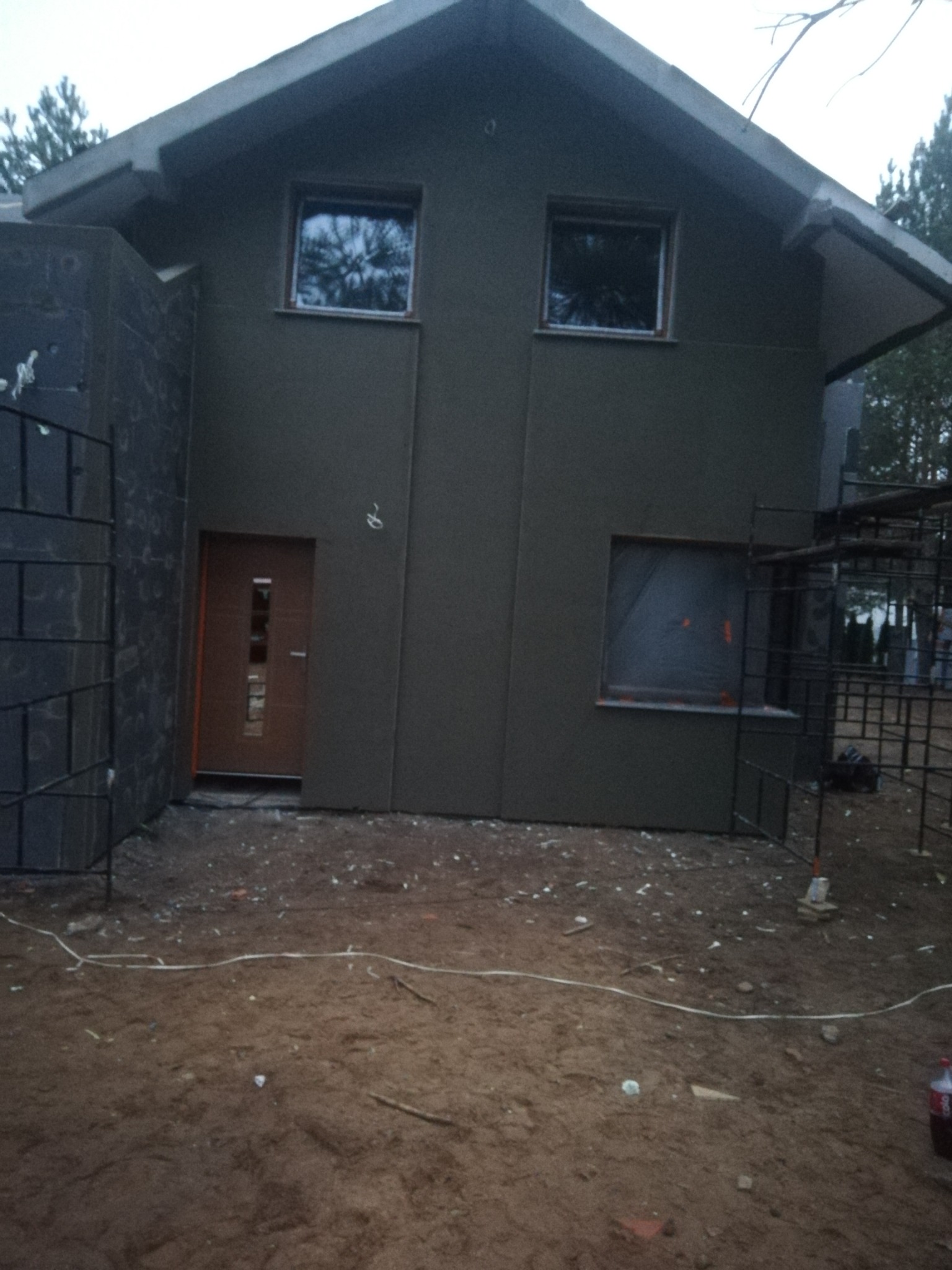 Surowy stan elewacji domu jednorodzinnego w trakcie budowy, z oknami i drzwiami, widoczne rusztowania i nierówny teren.