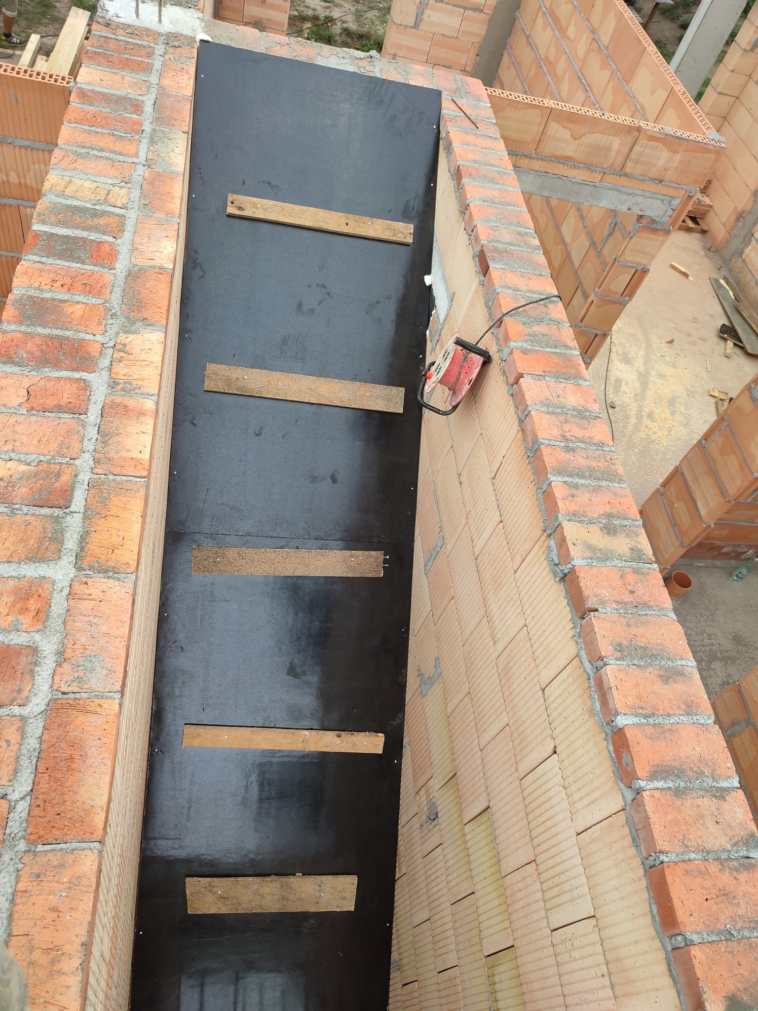 Wnętrze budowanego komina z czerwonej cegły ceramicznej, zabezpieczonego czarną folią i drewnianymi rozpórkami, widok z góry.