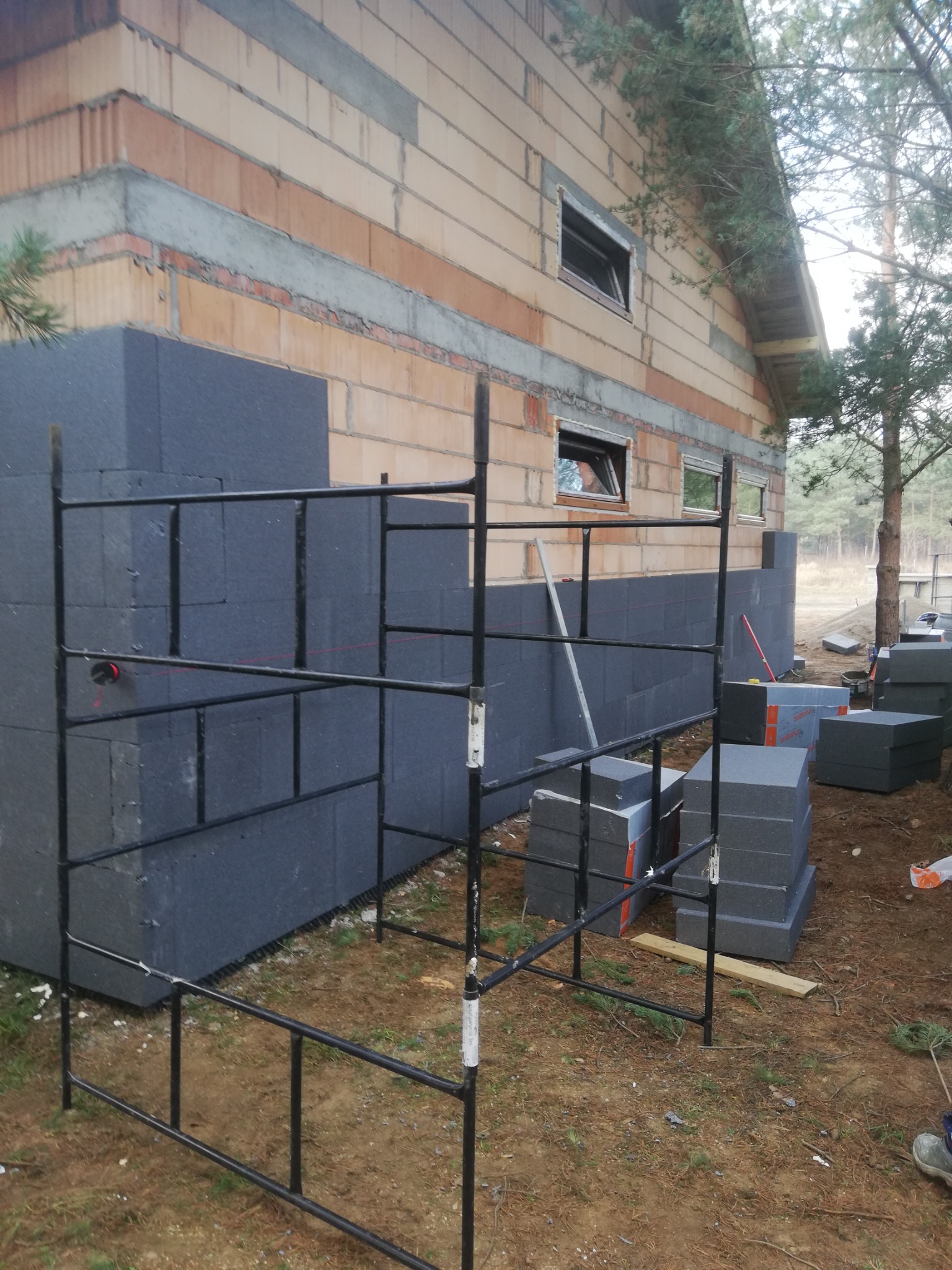 Ocieplanie ścian zewnętrznych domu jednorodzinnego szarym styropianem grafitowym, widoczne rusztowanie i ułożone bloki izolacyjne.