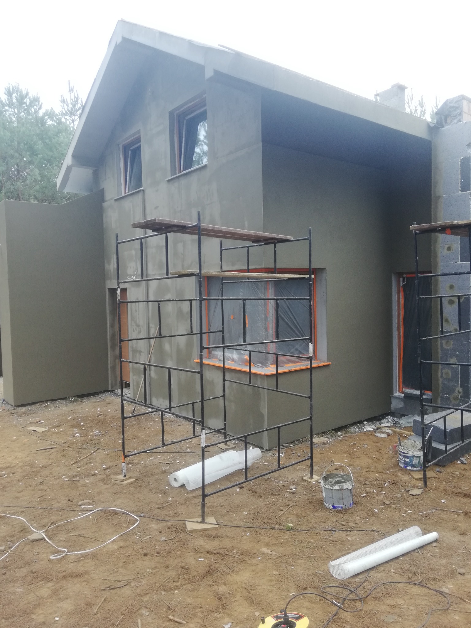 Dwukondygnacyjny dom w trakcie ocieplania i wykańczania elewacji, z widocznymi rusztowaniami, zabezpieczonymi oknami i fragmentami izolacji na ścianach.