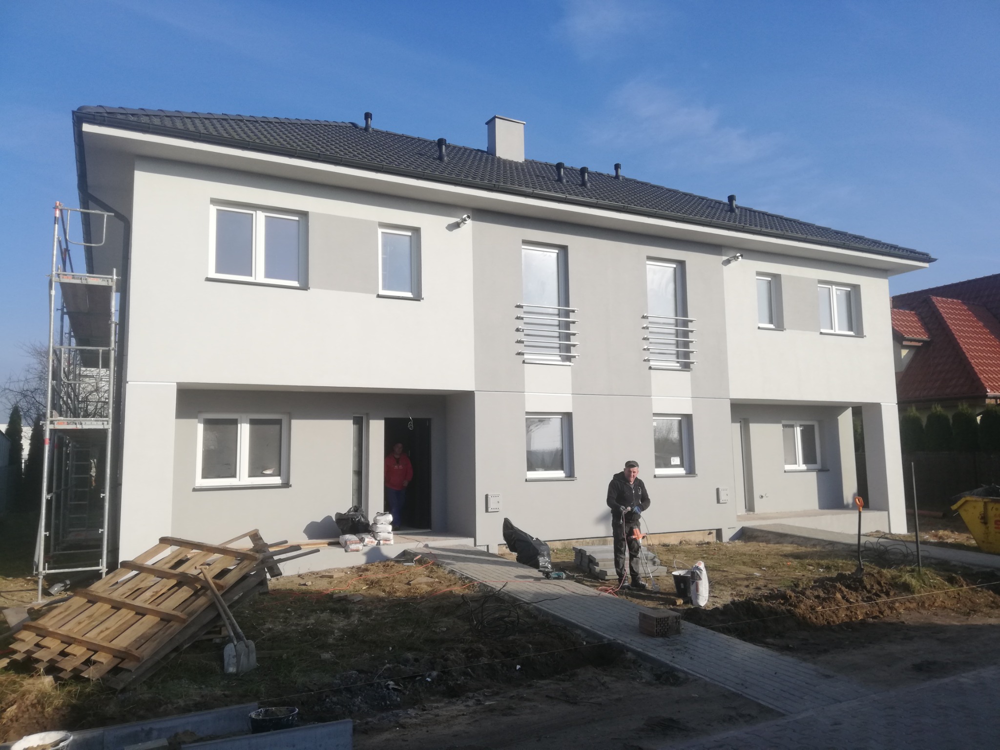 Dwukondygnacyjny budynek w trakcie wykończenia elewacji, z robotnikiem używającym narzędzia do obróbki kamienia na chodniku z kostki, palety i rusztowania obok budynku.