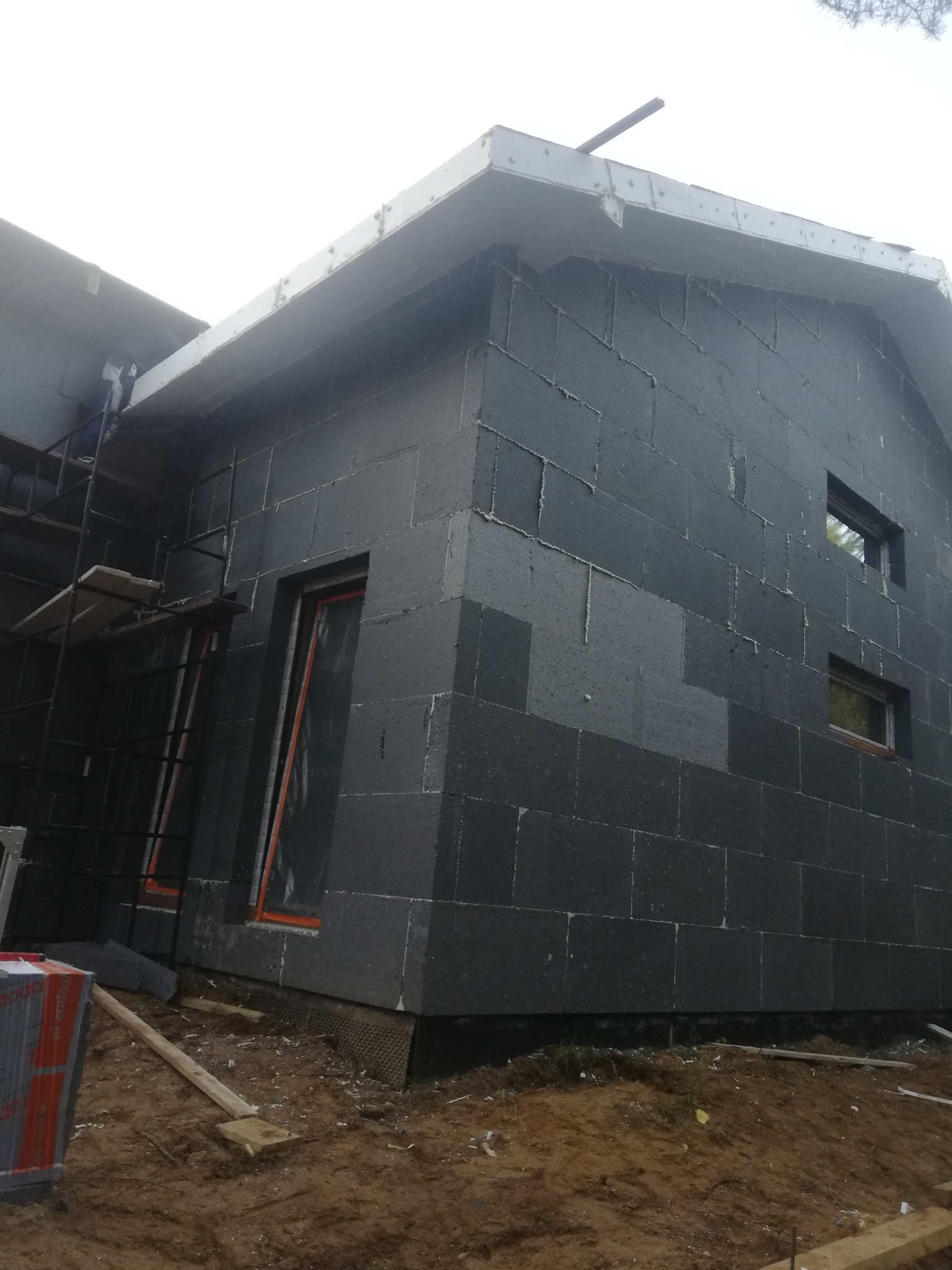 Budowa domu z szarych bloczków betonowych, widoczne okna w folii ochronnej, rusztowanie i materiały budowlane na ziemi.