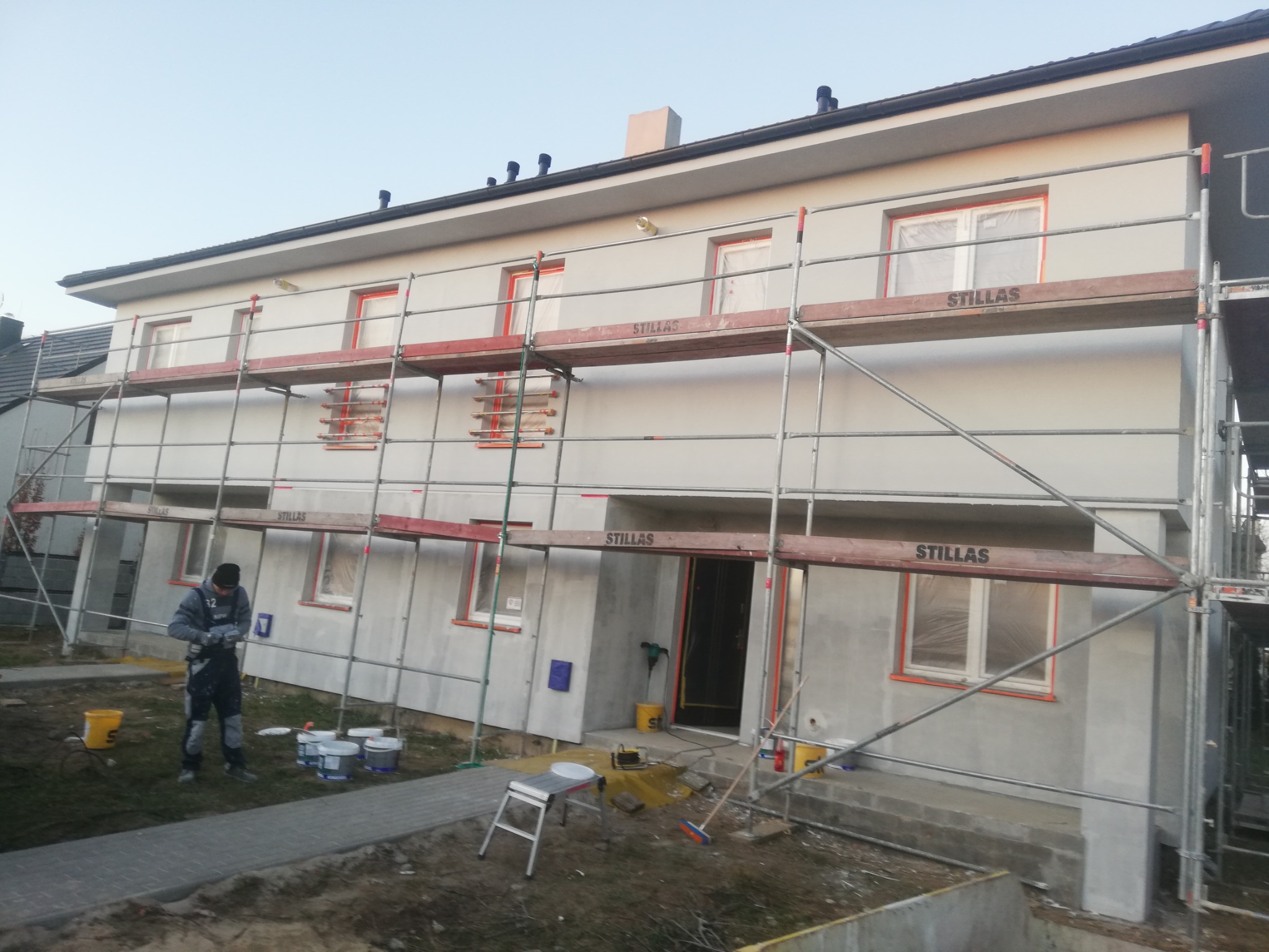 Dwukondygnacyjny dom w trakcie ocieplania, z widocznymi rusztowaniami, oknami zabezpieczonymi folią i pracownikiem przygotowującym się do pracy, z kilkoma puszkami farby obok.