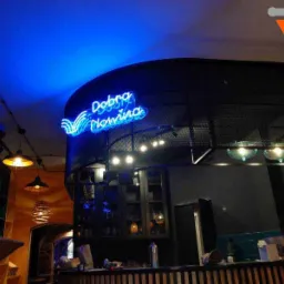Niebieski neonowy napis 'Dobra Nowina' w restauracji, oświetlenie sufitowe z wiszącymi lampami w stylu industrialnym, widok z dołu na bar.