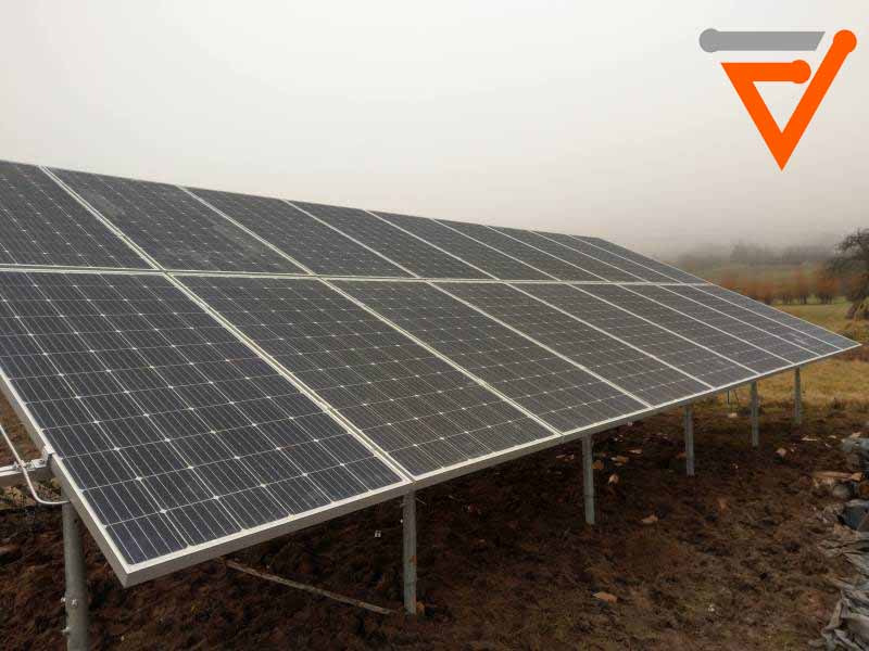 Instalacja fotowoltaiczna na gruncie, widoczne panele ustawione pod kątem, zamontowane na metalowych podporach. Tło stanowią drzewa we mgle.