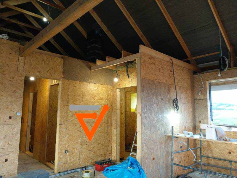 Wnętrze domu w trakcie budowy, widoczne ściany z płyt OSB, drewniana konstrukcja dachu, prowizoryczne oświetlenie i przewody elektryczne wystające ze ścian.