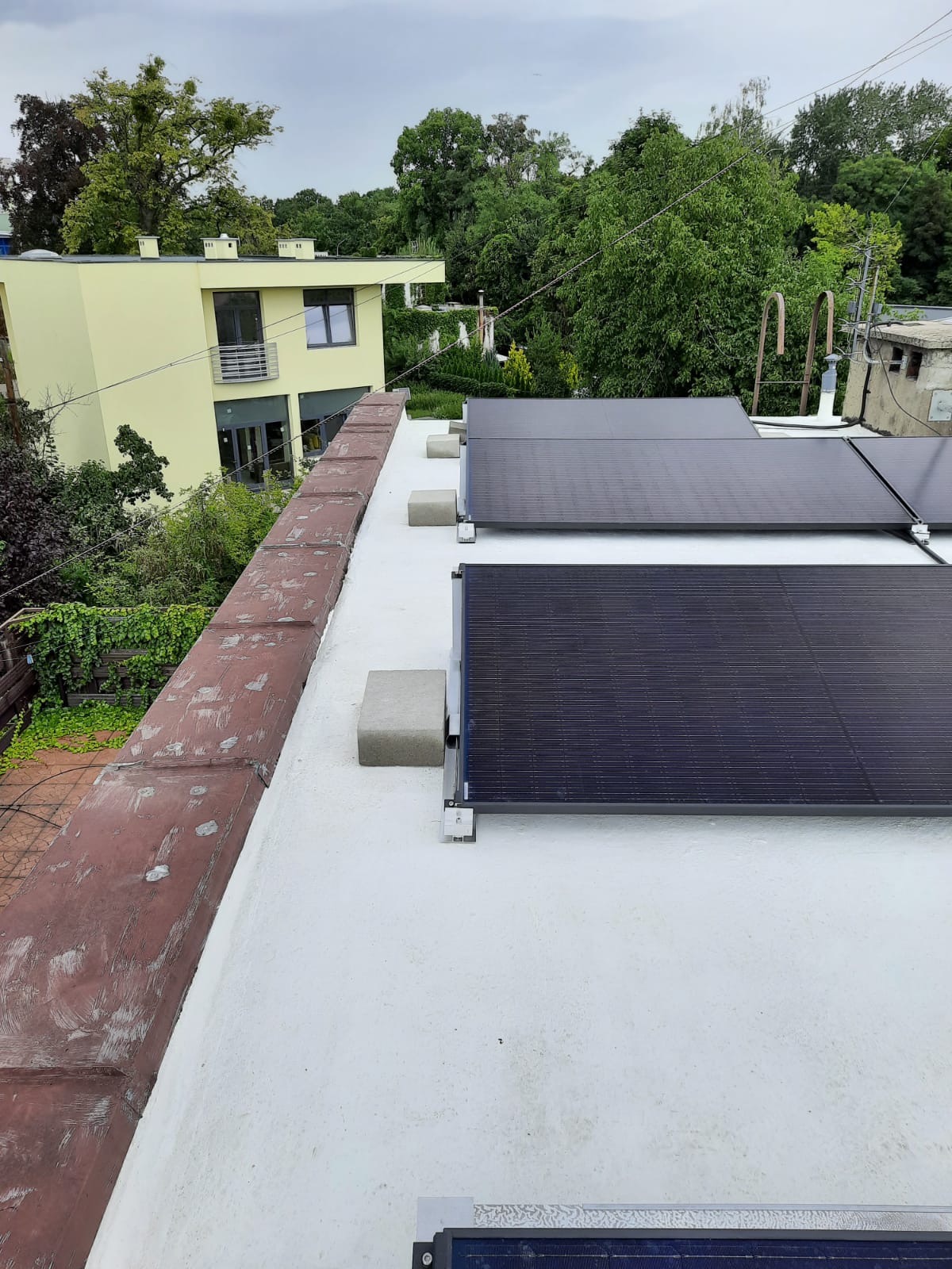 Panele słoneczne zamontowane na płaskim, białym dachu z widocznymi betonowymi blokami obciążającymi konstrukcję, w tle budynek i zieleń drzew.