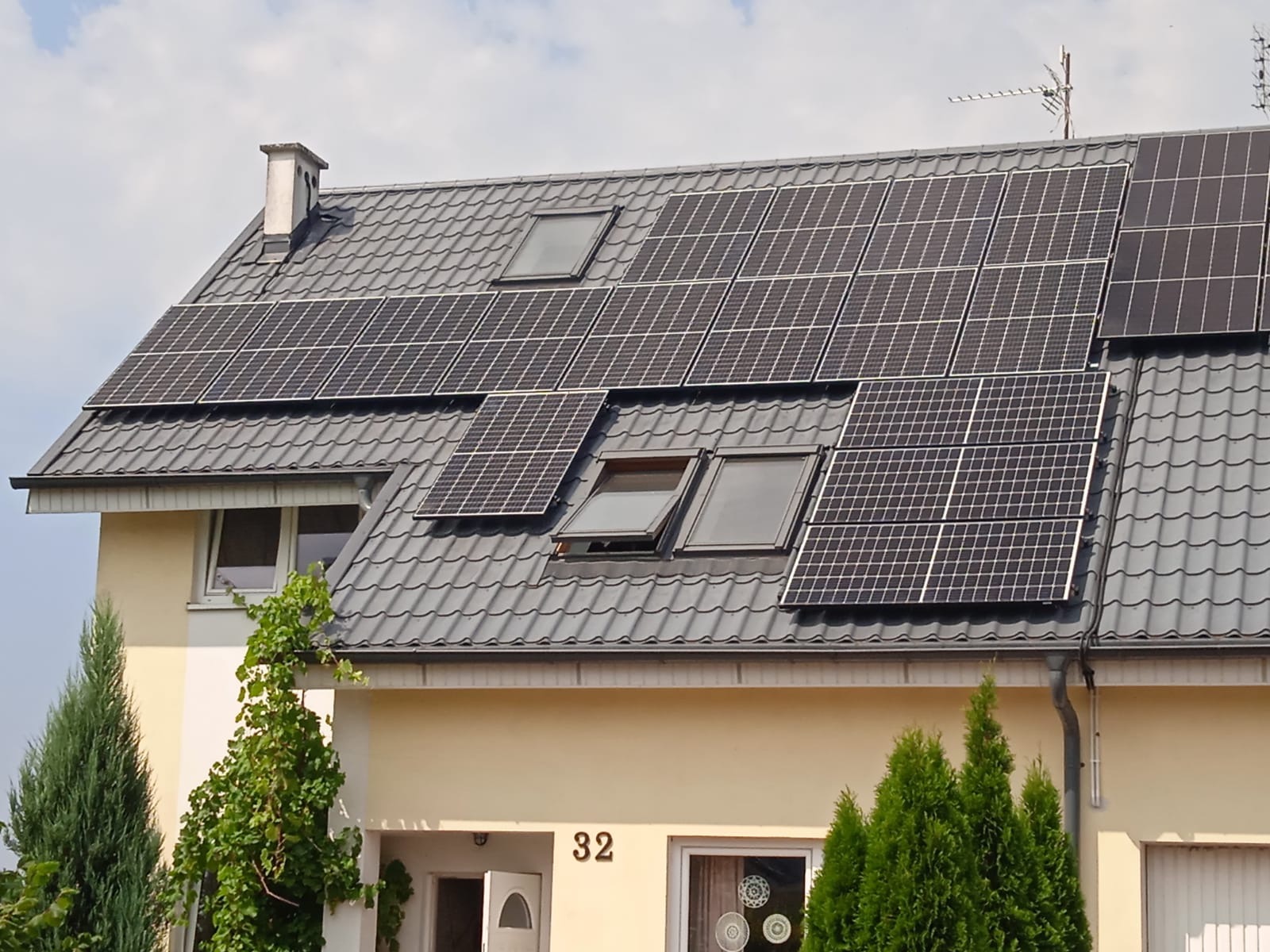 Panele słoneczne na dachu domu jednorodzinnego z szarą dachówką, widoczne okna dachowe i antena telewizyjna.