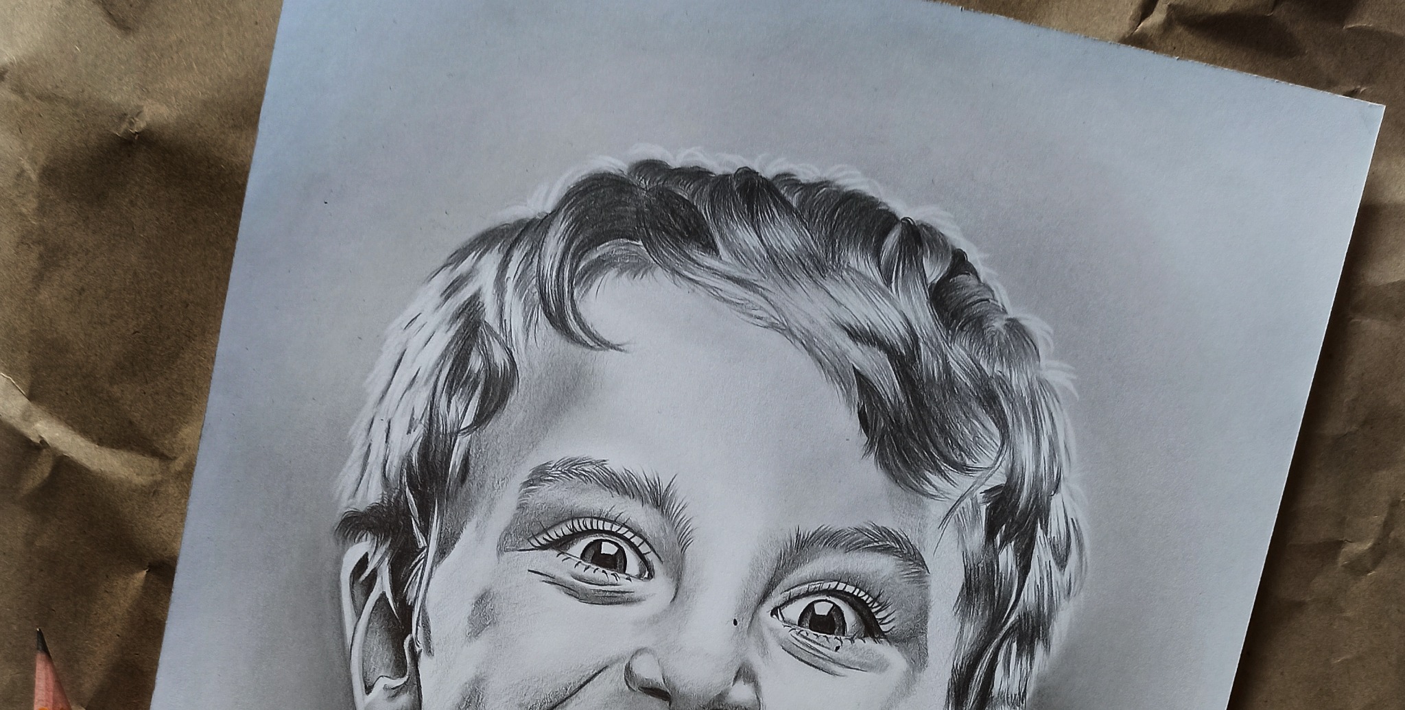 Portret chłopca narysowany ołówkiem, leżący na brązowym papierze, z widocznym ołówkiem w lewym dolnym rogu.