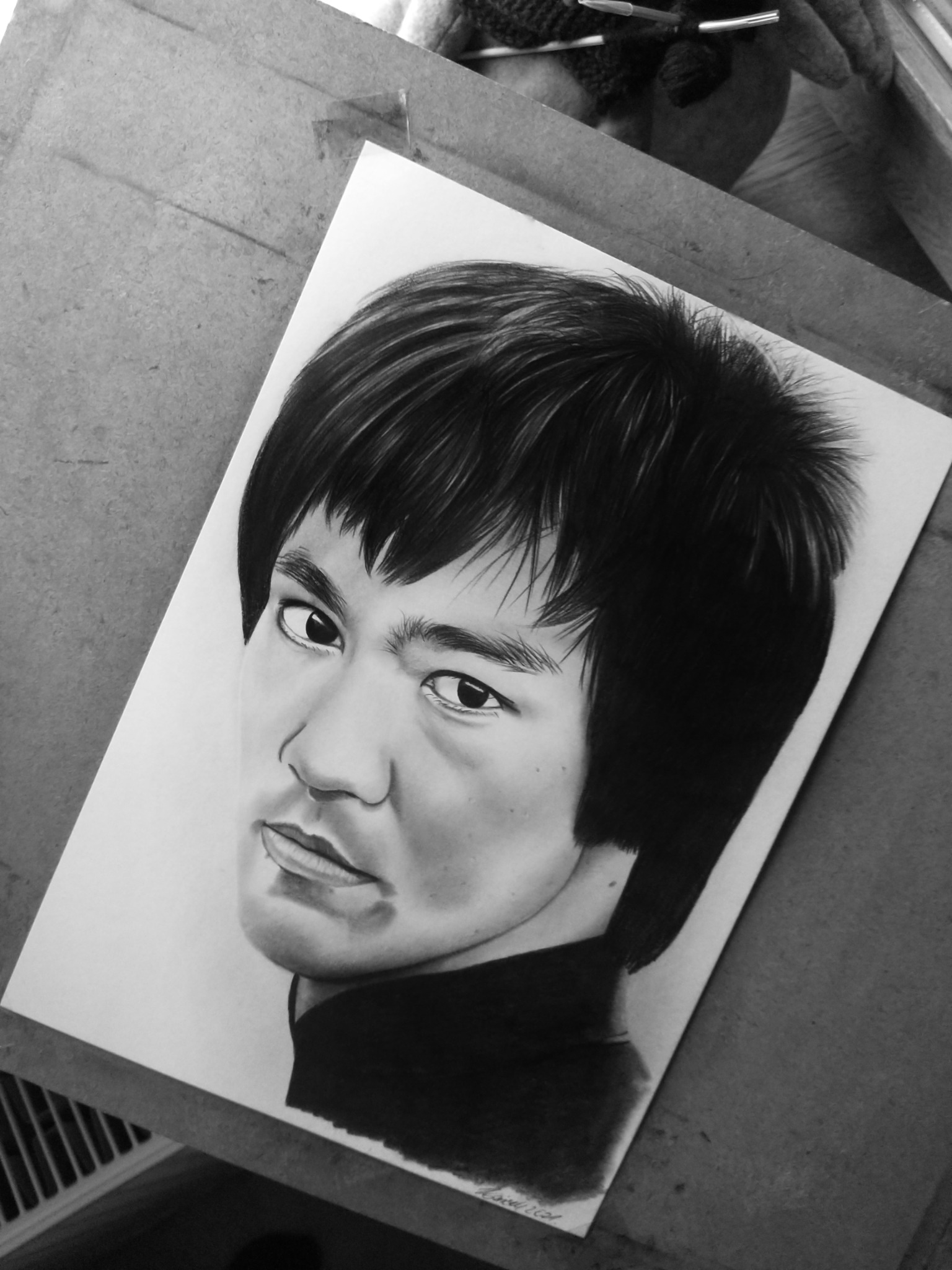 Czarno-biały portret Bruce'a Lee narysowany ołówkiem, leżący na biurku obok przyborów do rysowania.