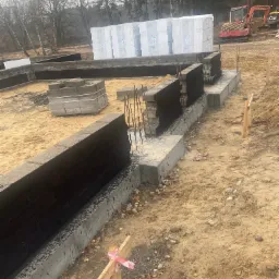 Wykopane fundamenty domu w trakcie budowy, z izolacją przeciwwilgociową, widoczne zbrojenie, palety z materiałami budowlanymi i maszyny budowlane w tle, teren piaszczysty.