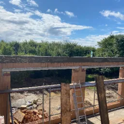 Wznoszenie ścian z bloczków ceramicznych, widoczne stalowe nadproże, rusztowanie i drabina, w tle zieleń drzew pod błękitnym niebem z chmurami.