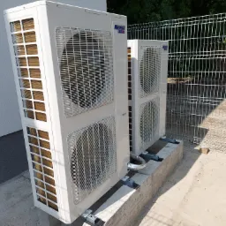 Dwie jednostki zewnętrzne klimatyzacji marki Gree, zamontowane na betonowym fundamencie przy ścianie budynku i ogrodzeniu z siatki.