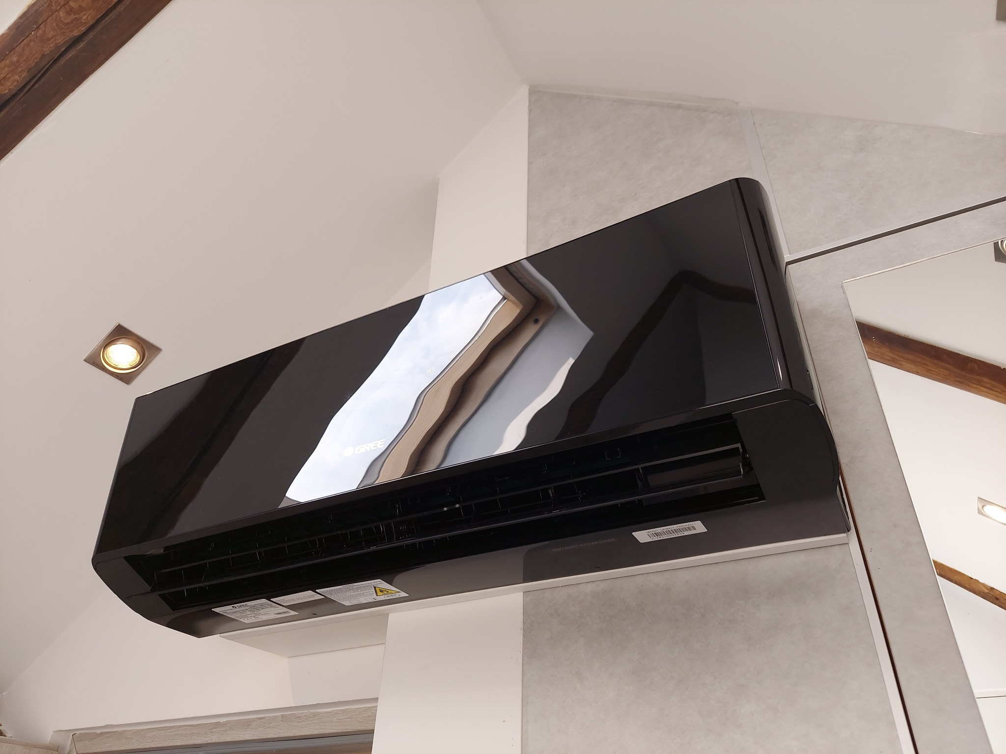 Czarny klimatyzator marki Gree zamontowany na ścianie w pomieszczeniu ze skośnym sufitem i drewnianymi belkami stropowymi.