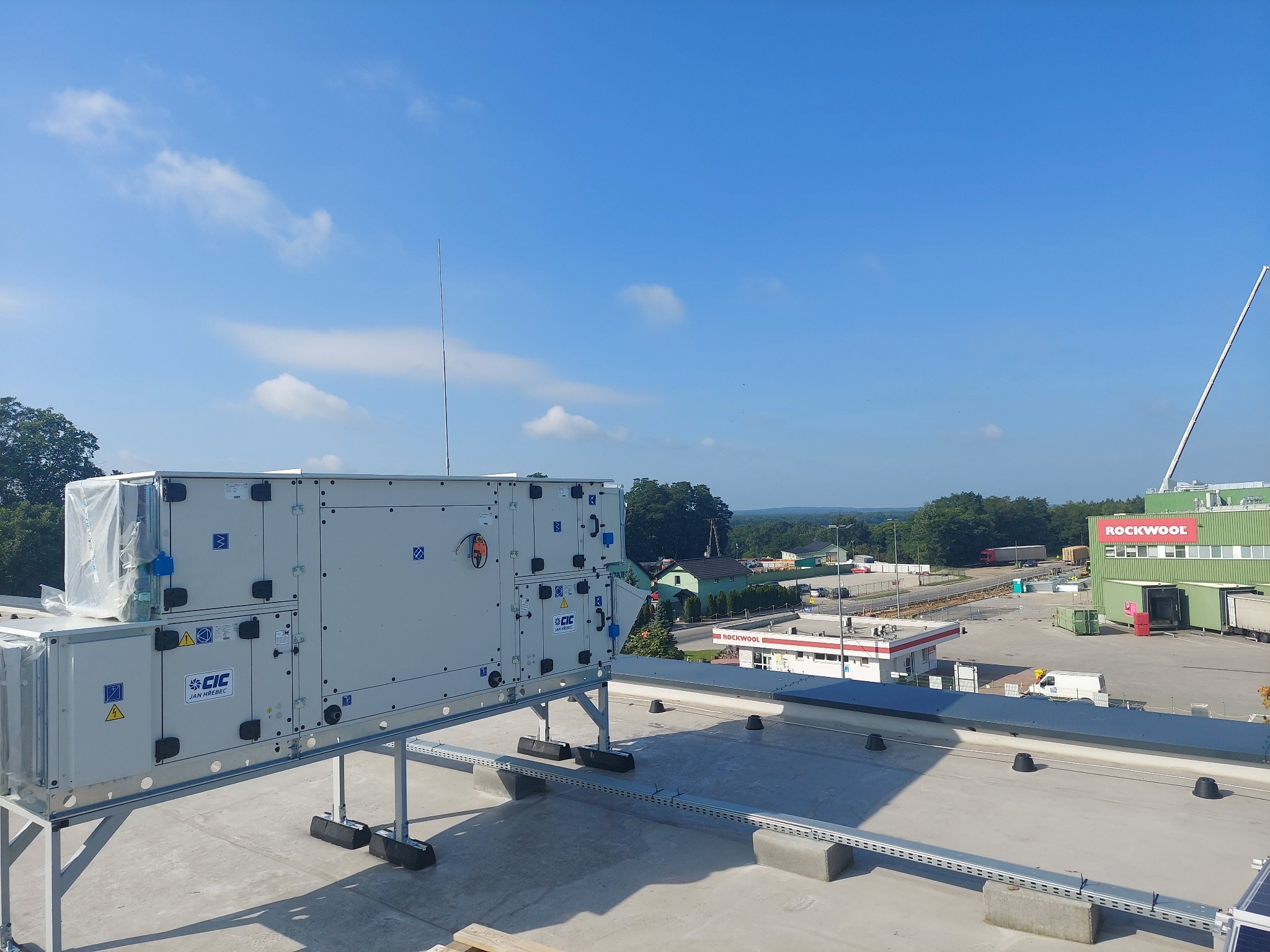 Centrala wentylacyjna na dachu budynku przemysłowego, z widocznym logo CIC Jan Hrebec i elementami instalacji w osłonach, na tle błękitnego nieba i zabudowań Rockwool.