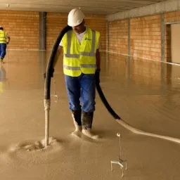 Pracownik w kasku i kamizelce wylewa płynną masę na podłogę z węża, w tle drugi pracownik i surowe ściany z cegły.
