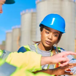 Inżynierowie w kaskach analizują dane na tablecie na tle przemysłowych silosów, w jasnym świetle dnia.