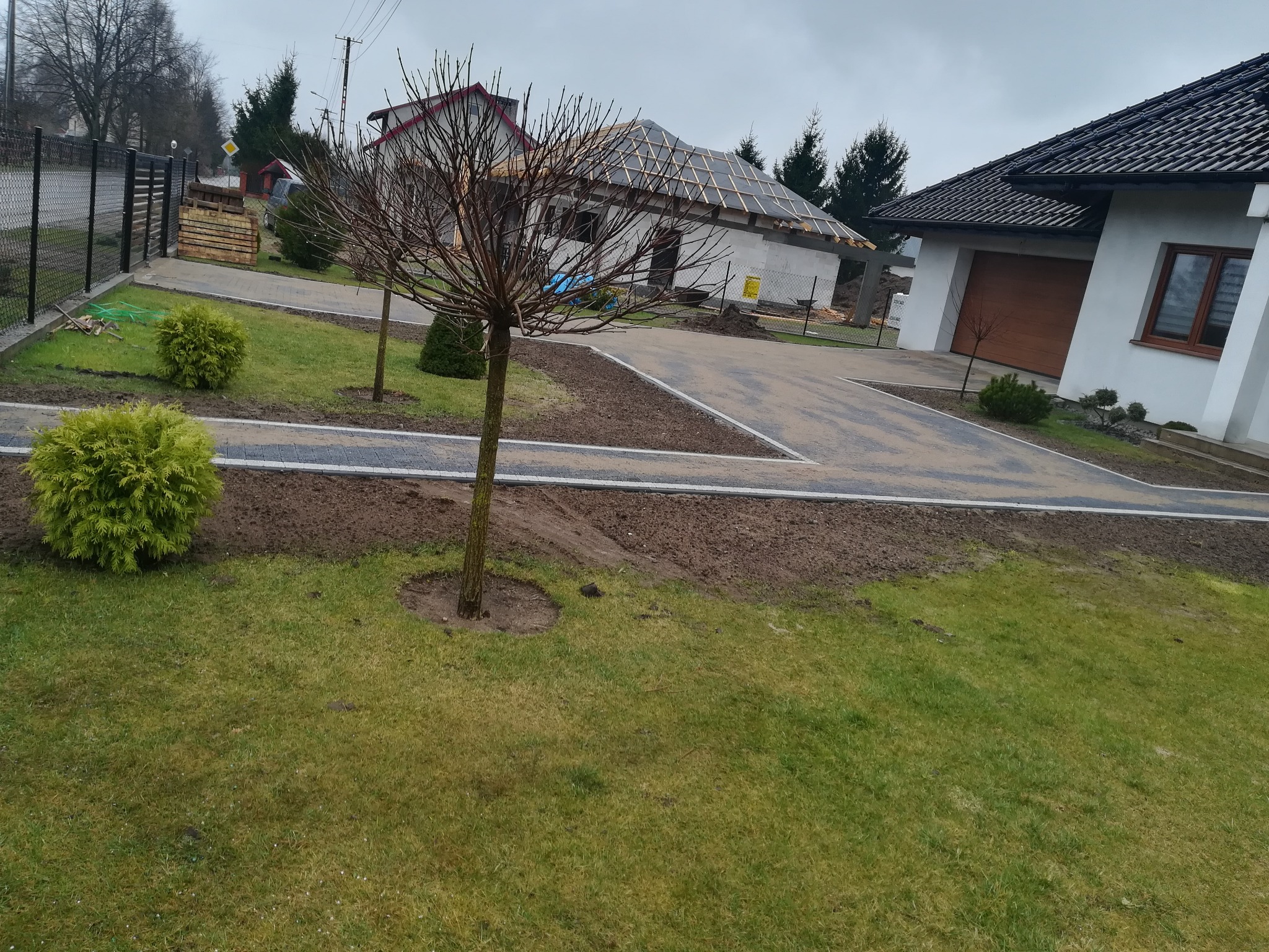 Ułożona nawierzchnia z kostki brukowej prowadząca do domu w trakcie budowy, widoczne elementy ogrodzenia i zagospodarowania terenu zielonego.