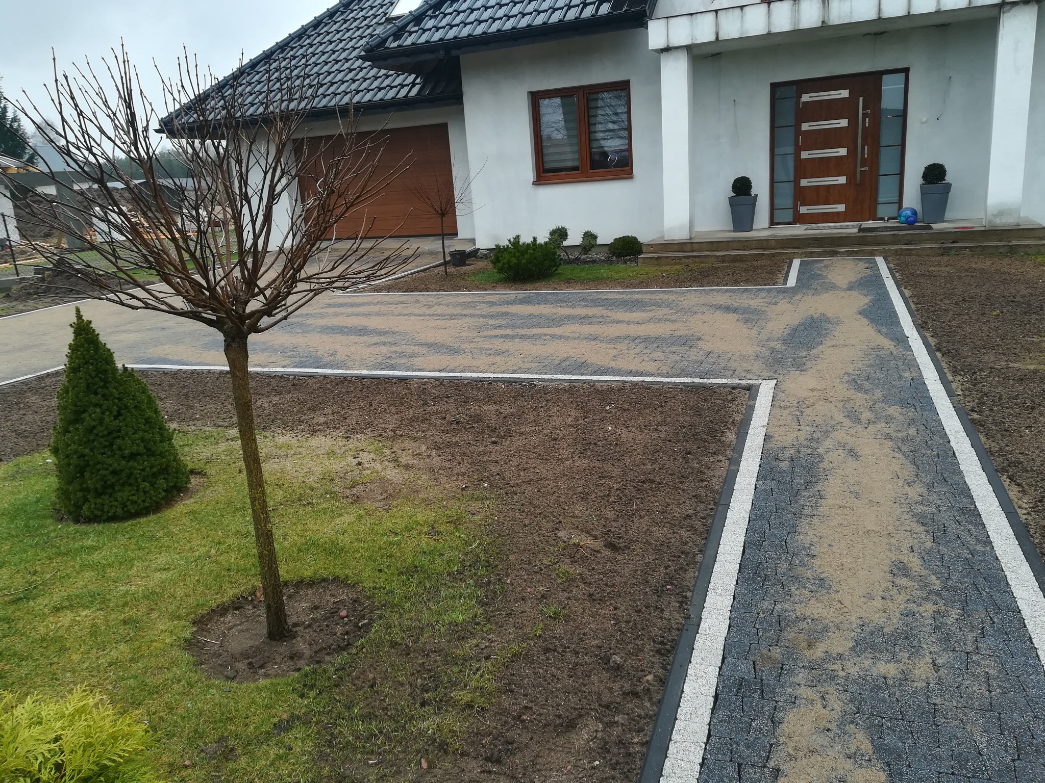 Nowo ułożony chodnik z szarej kostki brukowej z białymi obrzeżami prowadzi do nowoczesnego domu z brązowymi drzwiami i garażem, na trawniku rosną drzewka i krzewy.