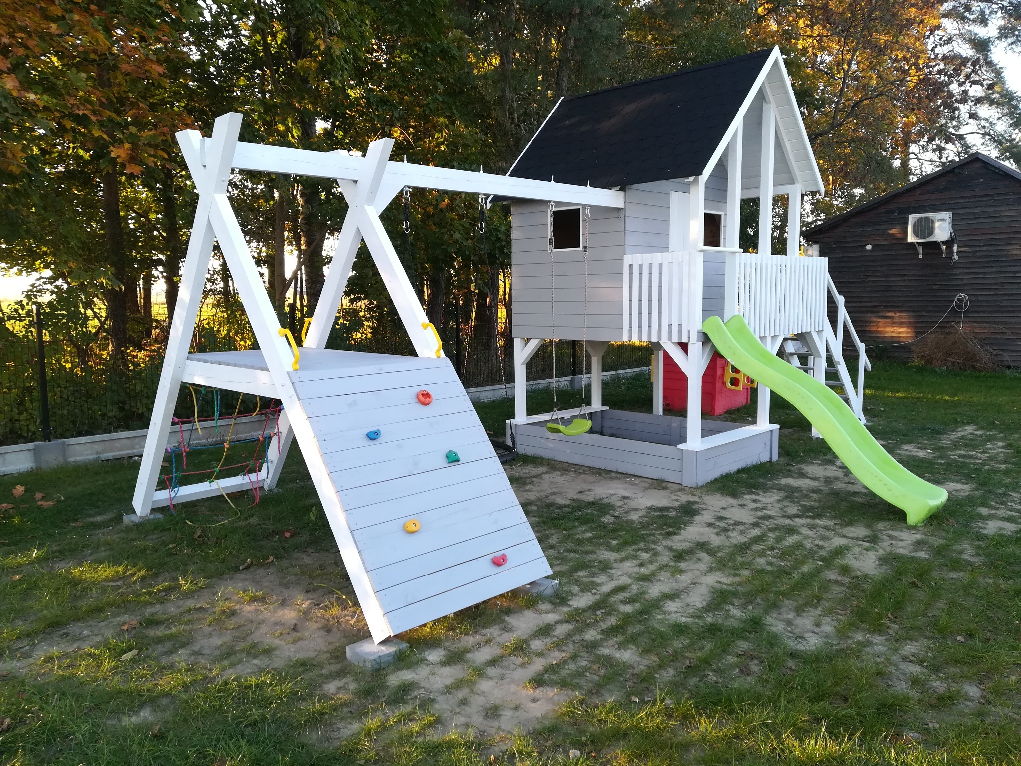 Kompleksowy plac zabaw z drewnianym domkiem na podeście, ścianką wspinaczkową z kolorowymi uchwytami, huśtawką i zieloną zjeżdżalnią na trawniku, w tle drzewa i budynek z klimatyzacją.