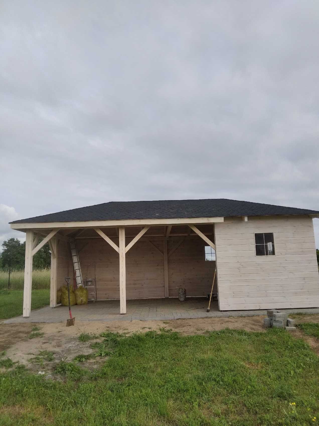 Drewniana altana z czarnym dachem z gontu bitumicznego, częściowo wykończona, z widoczną drabiną i workami w środku, położona na nowo ułożonej kostce brukowej.