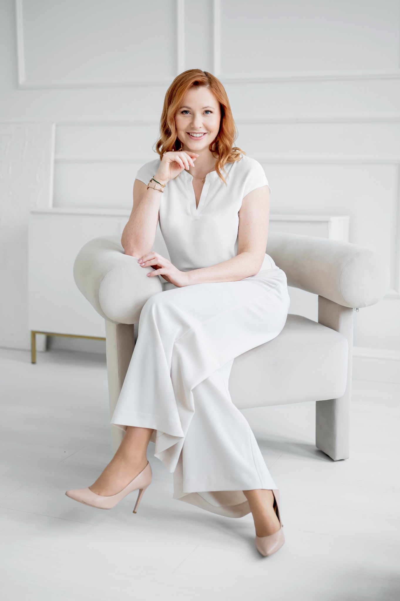 Uśmiechnięta kobieta z rudymi włosami, ubrana w biały strój, siedzi w beżowym fotelu na tle białej ściany. Prezentacja agenta nieruchomości.